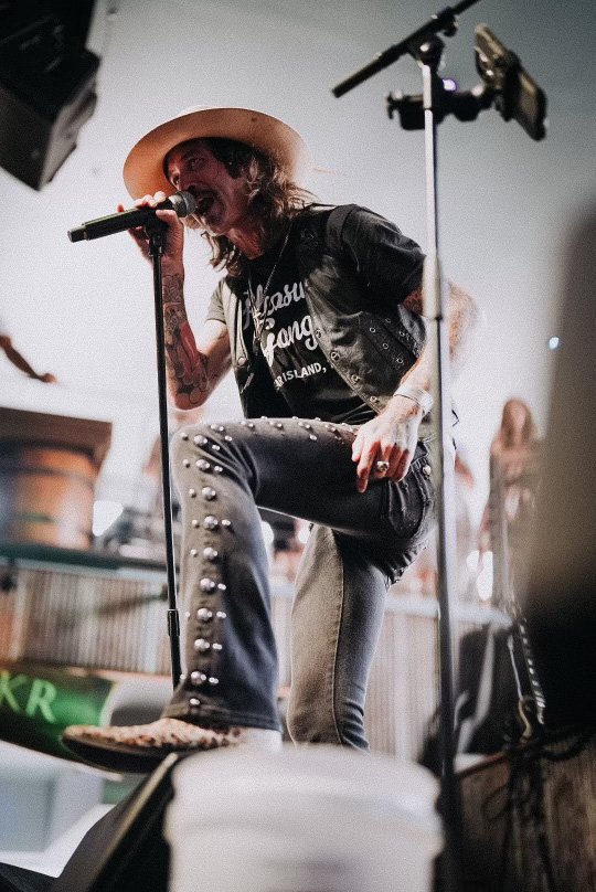 A male singer with long hair and tattoos wearing a wide-brimmed hat, black vest, and jeans with metal studs, performing on stage with a microphone.