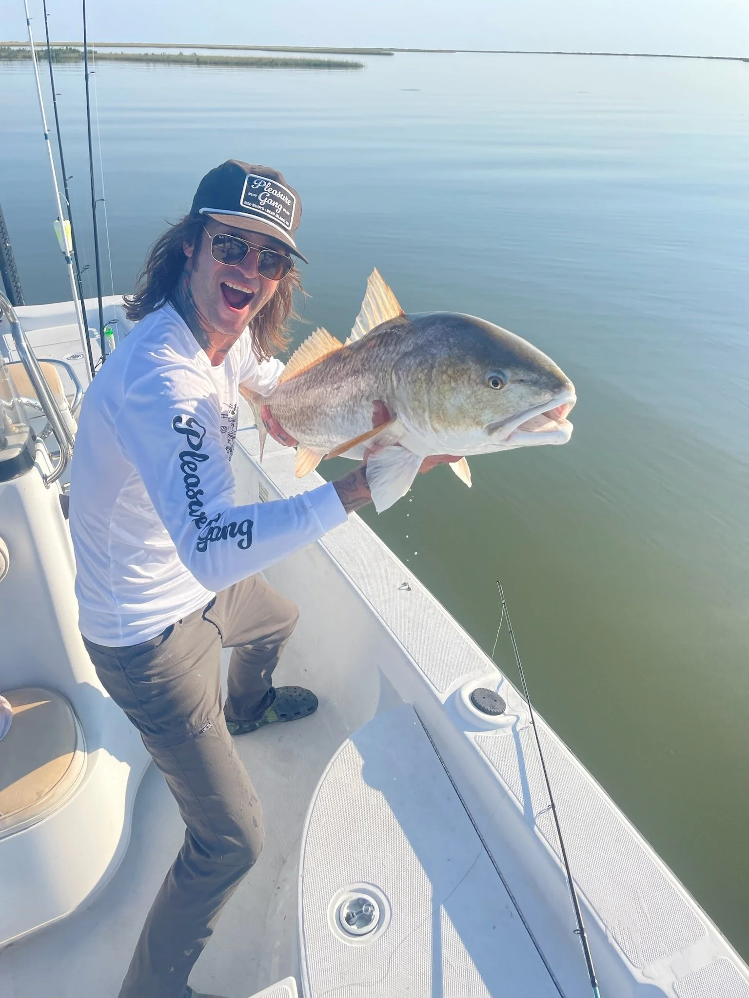 A smiling man on a boat holding a large fish he caught, with fishing rods visible, water and distant shoreline in the background.
