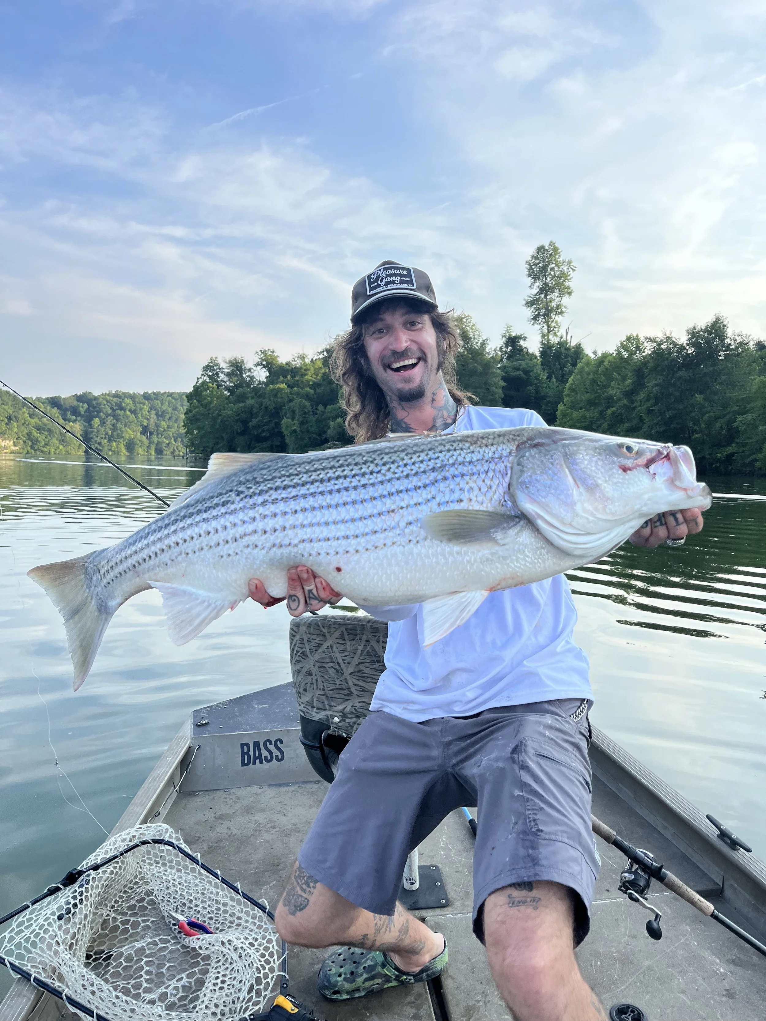 Man in a boat holding a large striped bass fish on a lake with green trees and blue sky in the background.