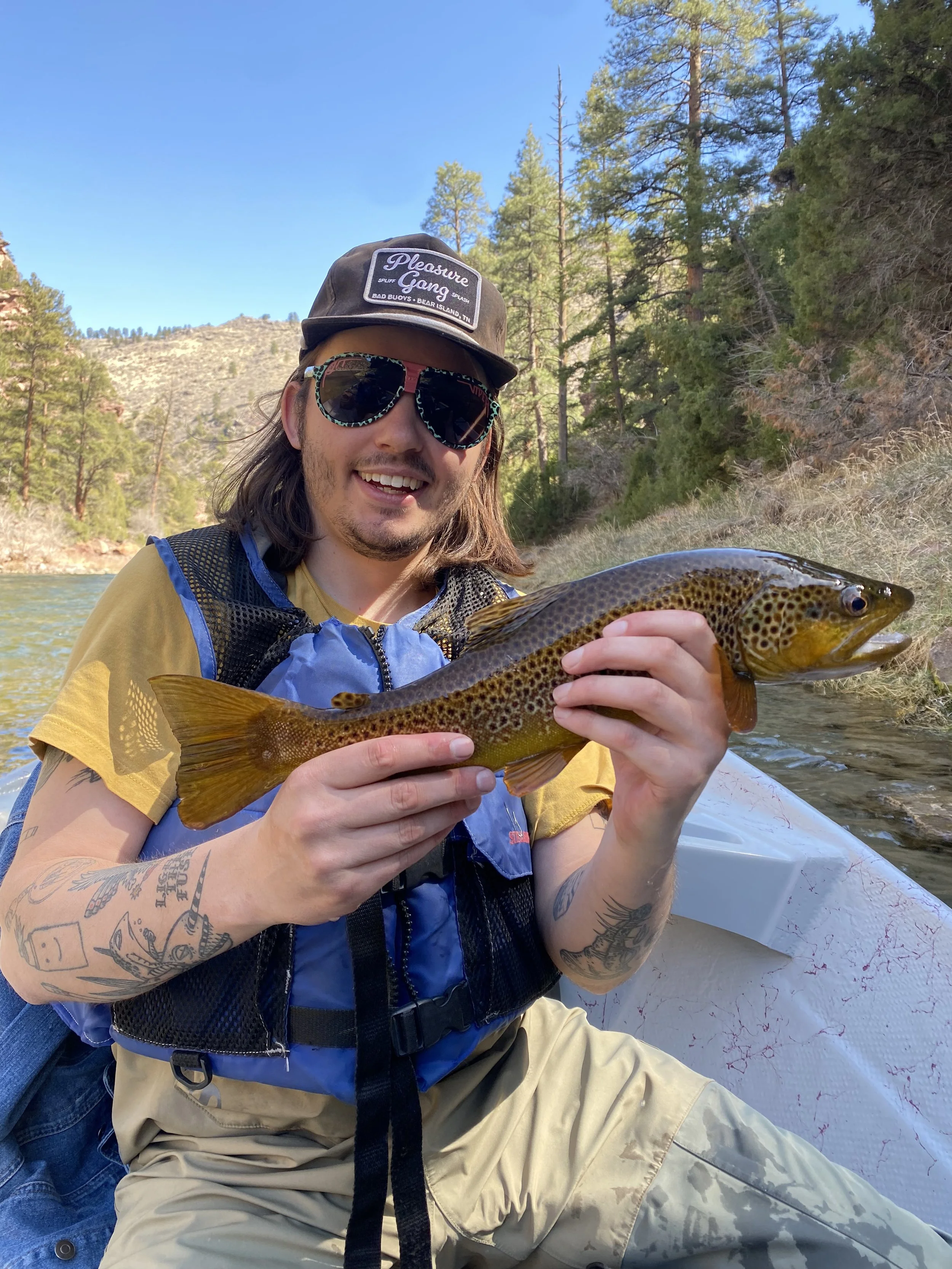 Man in sunglasses and a cap holding a large fish on a boat in a river surrounded by trees and mountains.