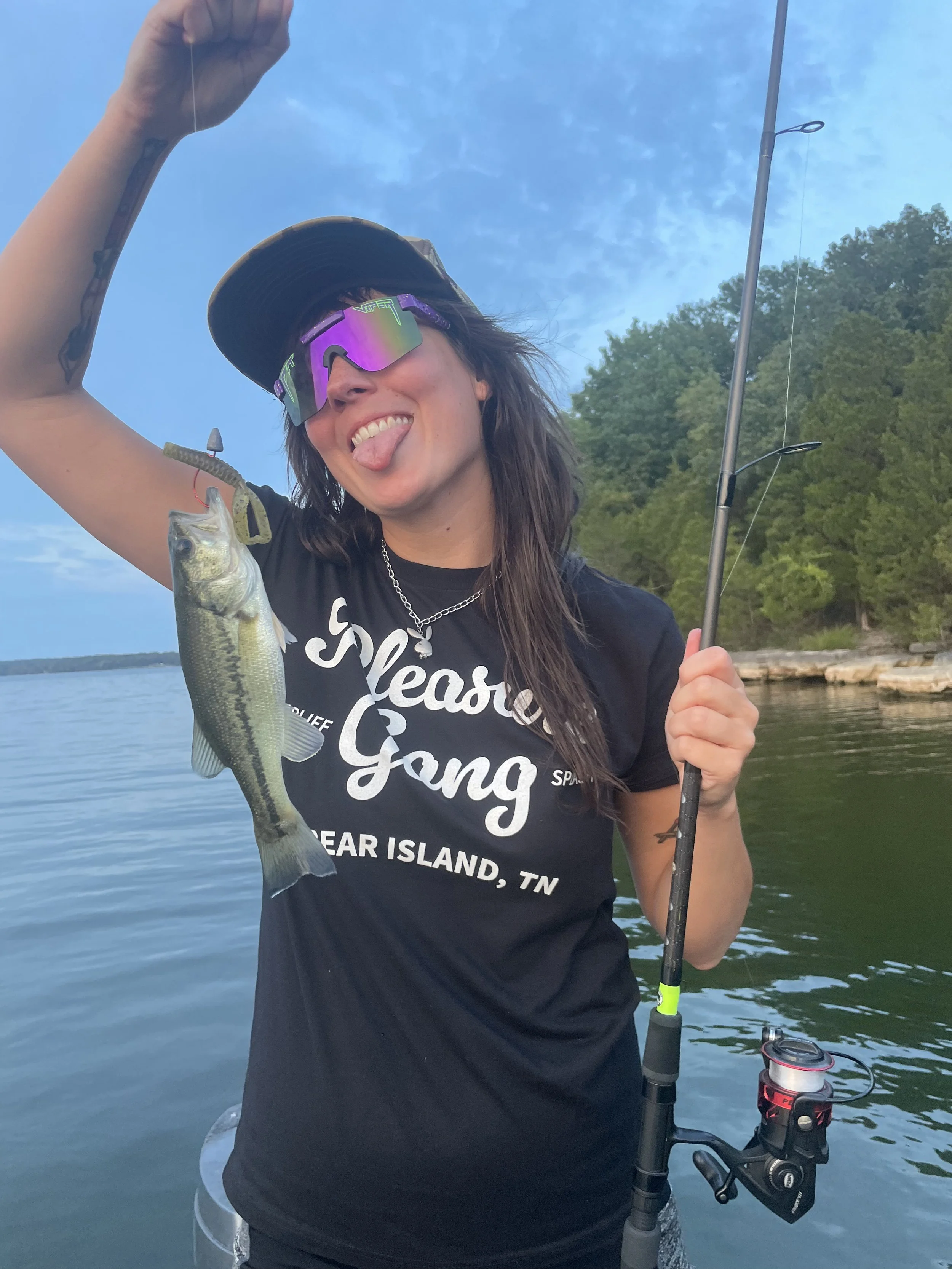 A woman on a boat holding a fishing rod with a large fish she caught, smiling and sticking her tongue out, wearing sunglasses, a cap, and a black t-shirt that says 'Sleek Gang' with 'GREAT ISLAND, TN' on it.