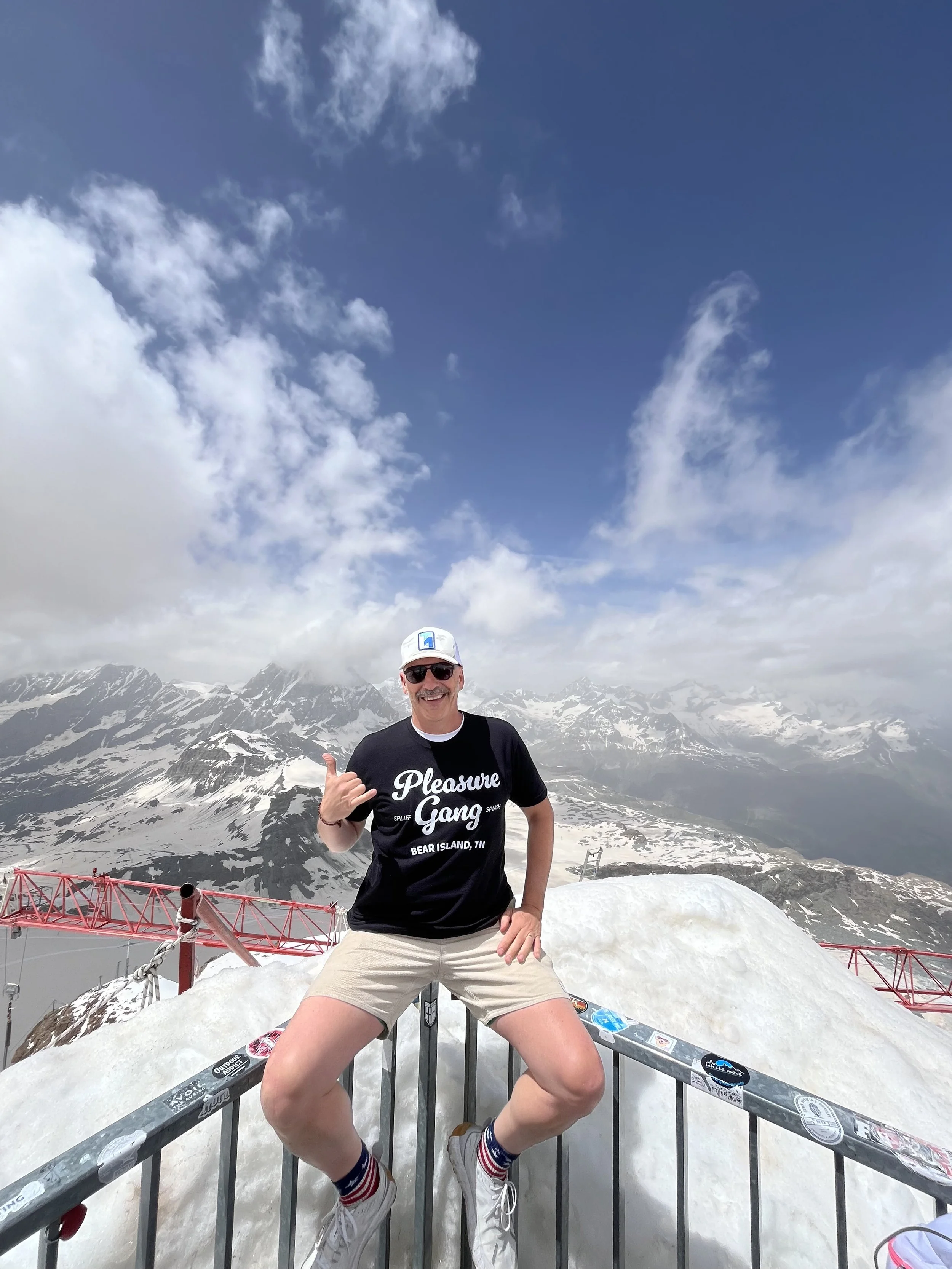 A man wearing sunglasses, a black t-shirt, beige shorts, and white sneakers with colorful striped socks is posing on a mountaintop with snow, surrounded by clouds and snow-capped peaks in the background.