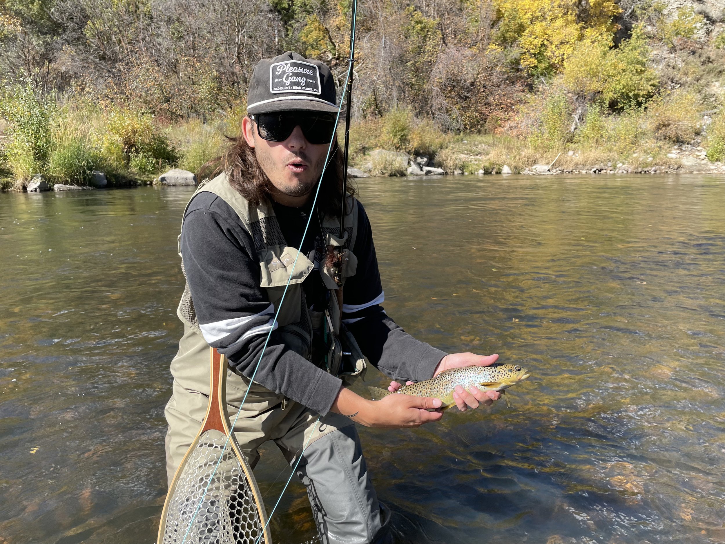 Man wearing sunglasses and hat holding a fish while standing in a river with fall foliage in the background.