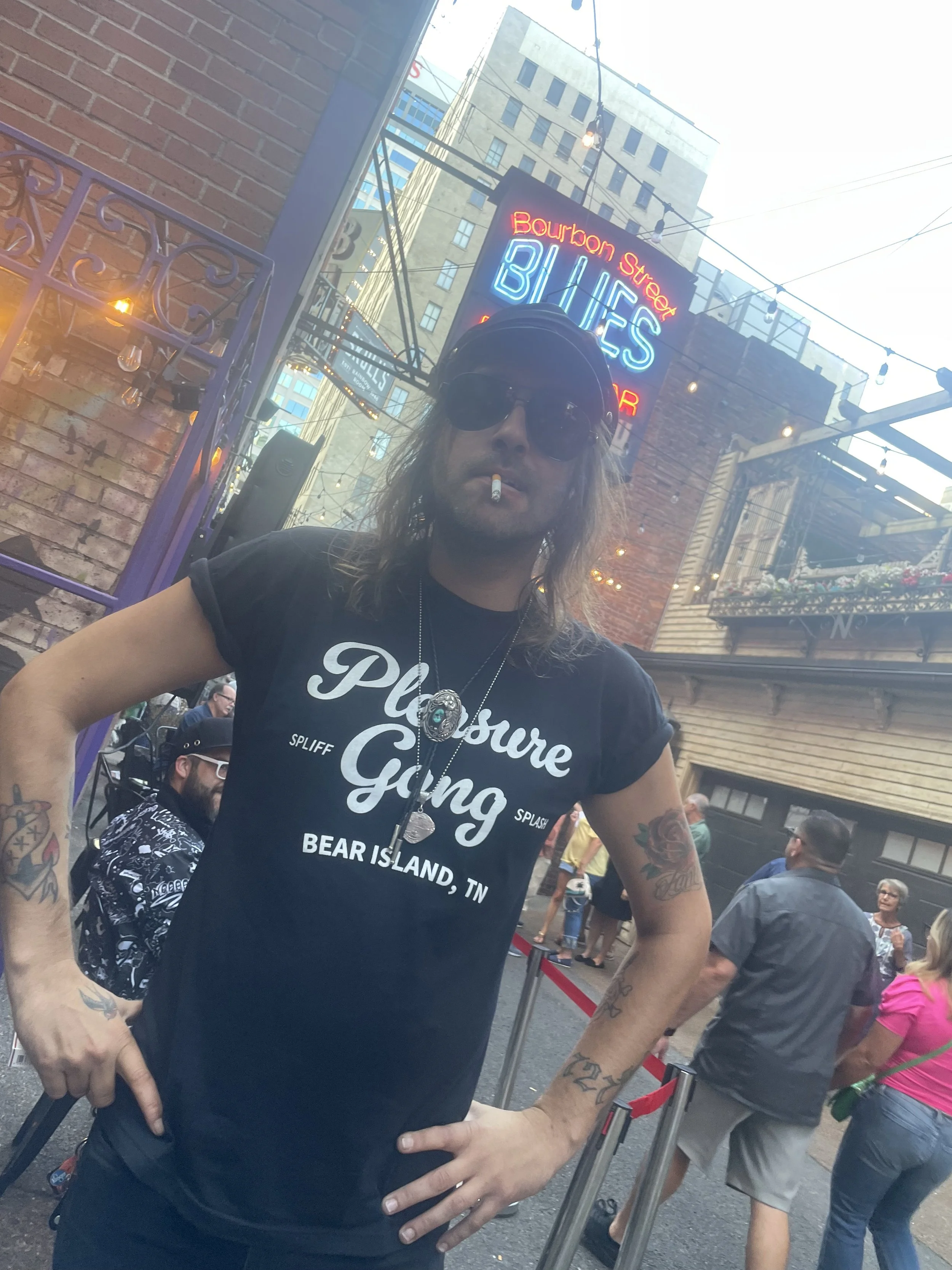 A man with long hair, wearing sunglasses, a beanie, and a black T-shirt that says 'Pleasure Ganging' with locations "Spliff," "Splash," and "Bear Island, TN". He has tattoos on both arms and a cigarette in his mouth. The background features a neon si