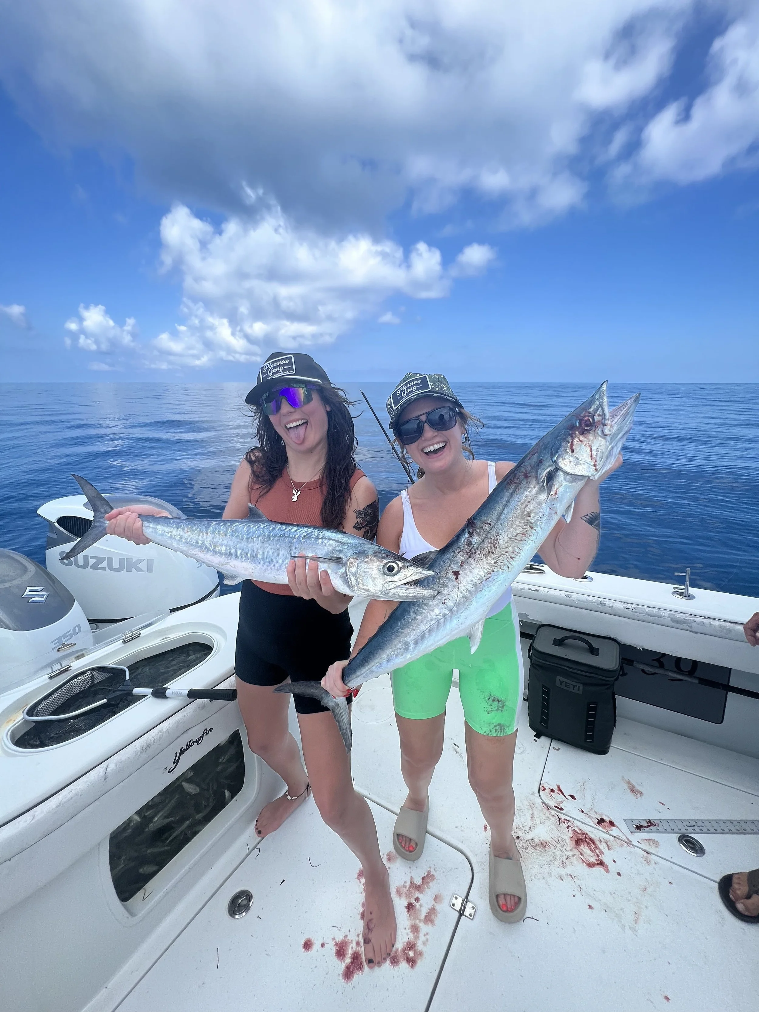 Two women on a boat holding large fish, smiling and posing for the camera, with the ocean and partly cloudy sky in the background.