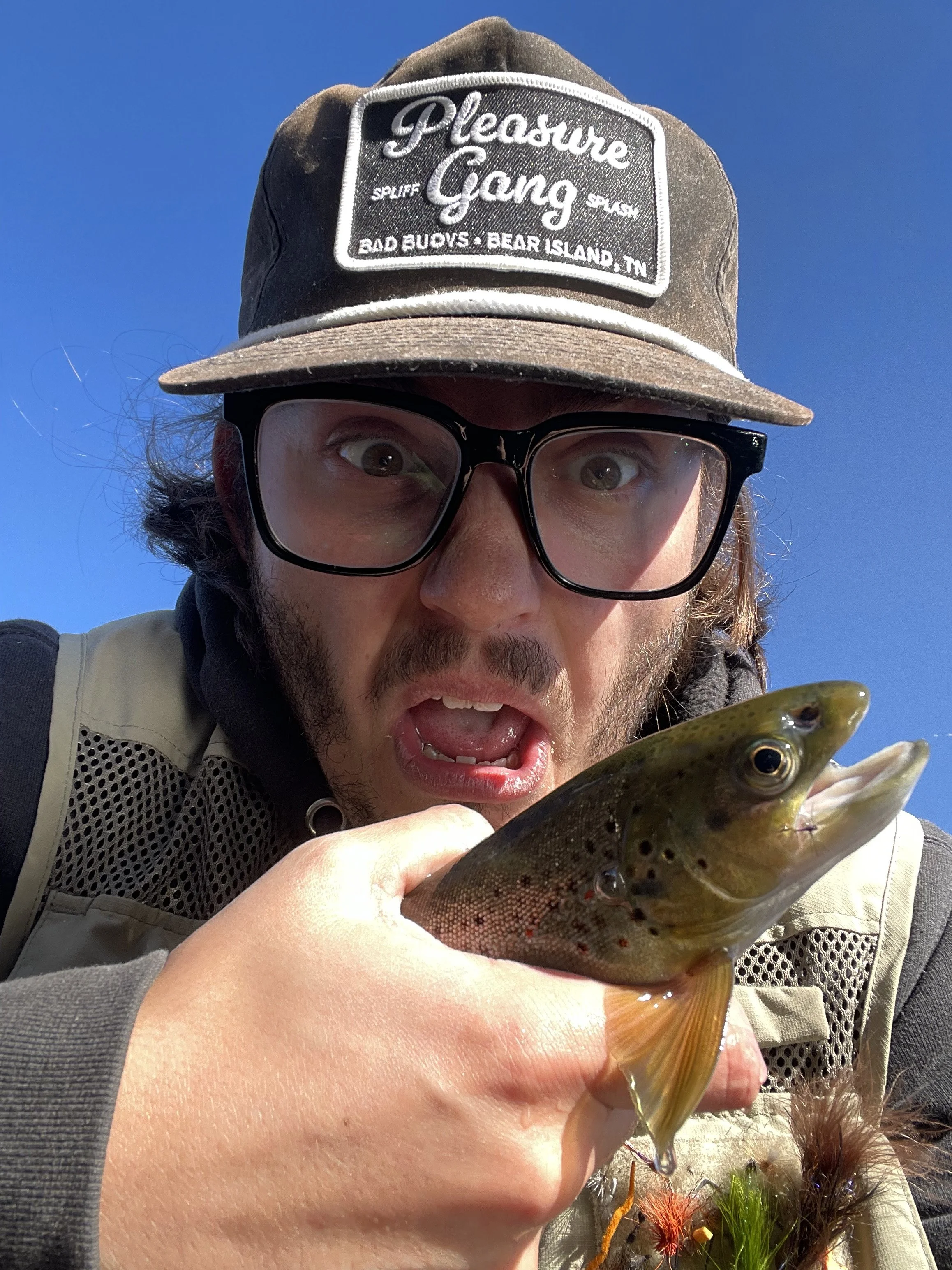A man with glasses, a cap, and a fishing vest holding a fish in front of a clear blue sky.