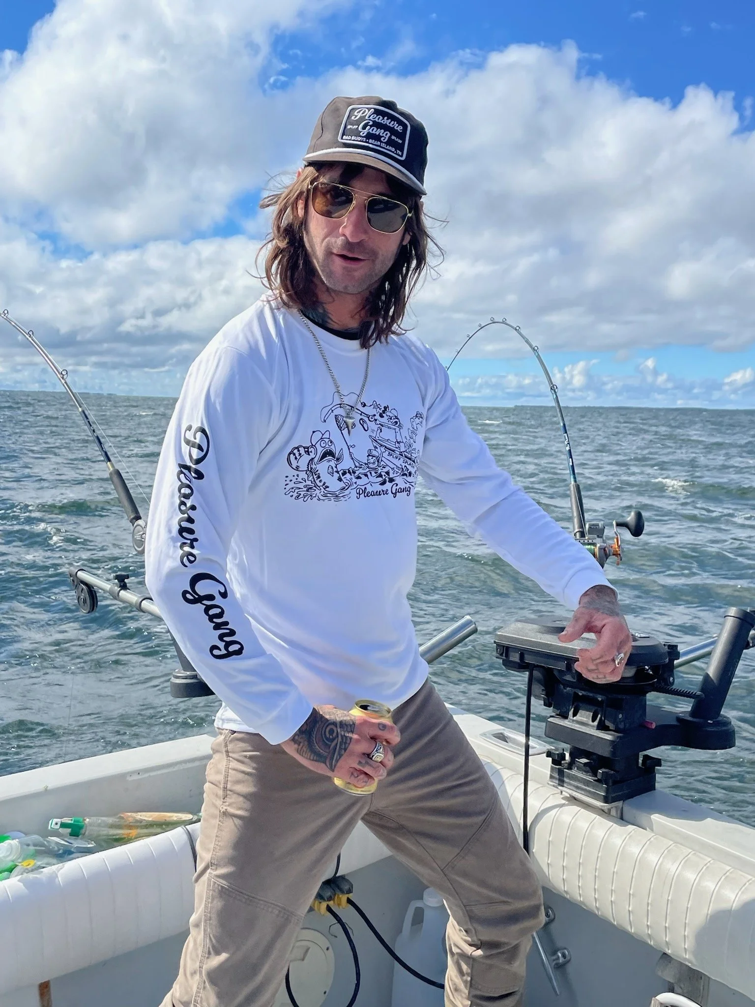 A man with long hair, wearing sunglasses, a baseball cap, and a white long-sleeve shirt with 'Pleasure Gang' written on the sleeve, standing on a boat with fishing rods, holding a drink can, in the ocean under a partly cloudy sky.