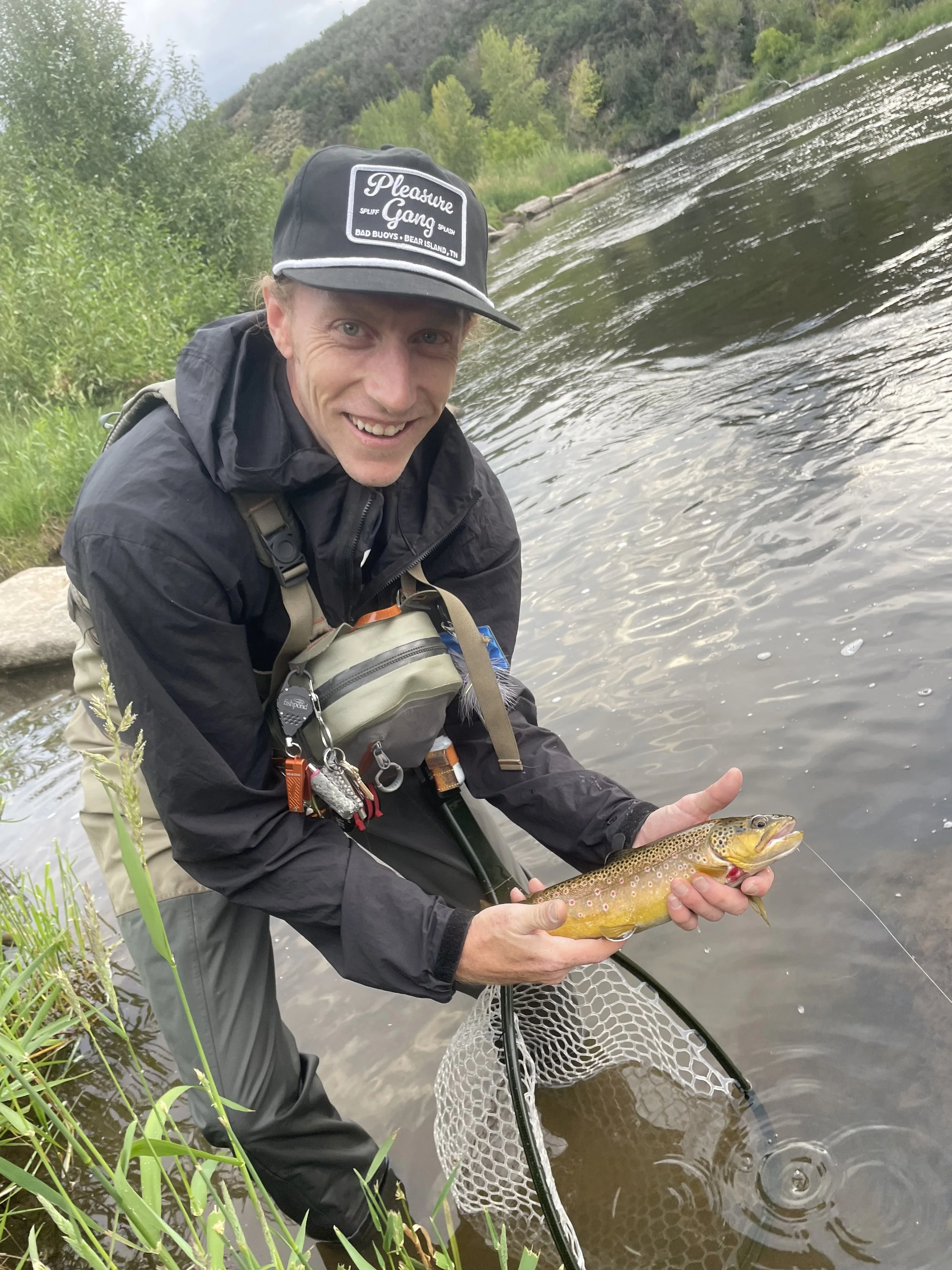Man in black jacket and cap holding a brown trout he caught near a riverbank with a fishing net in the water.