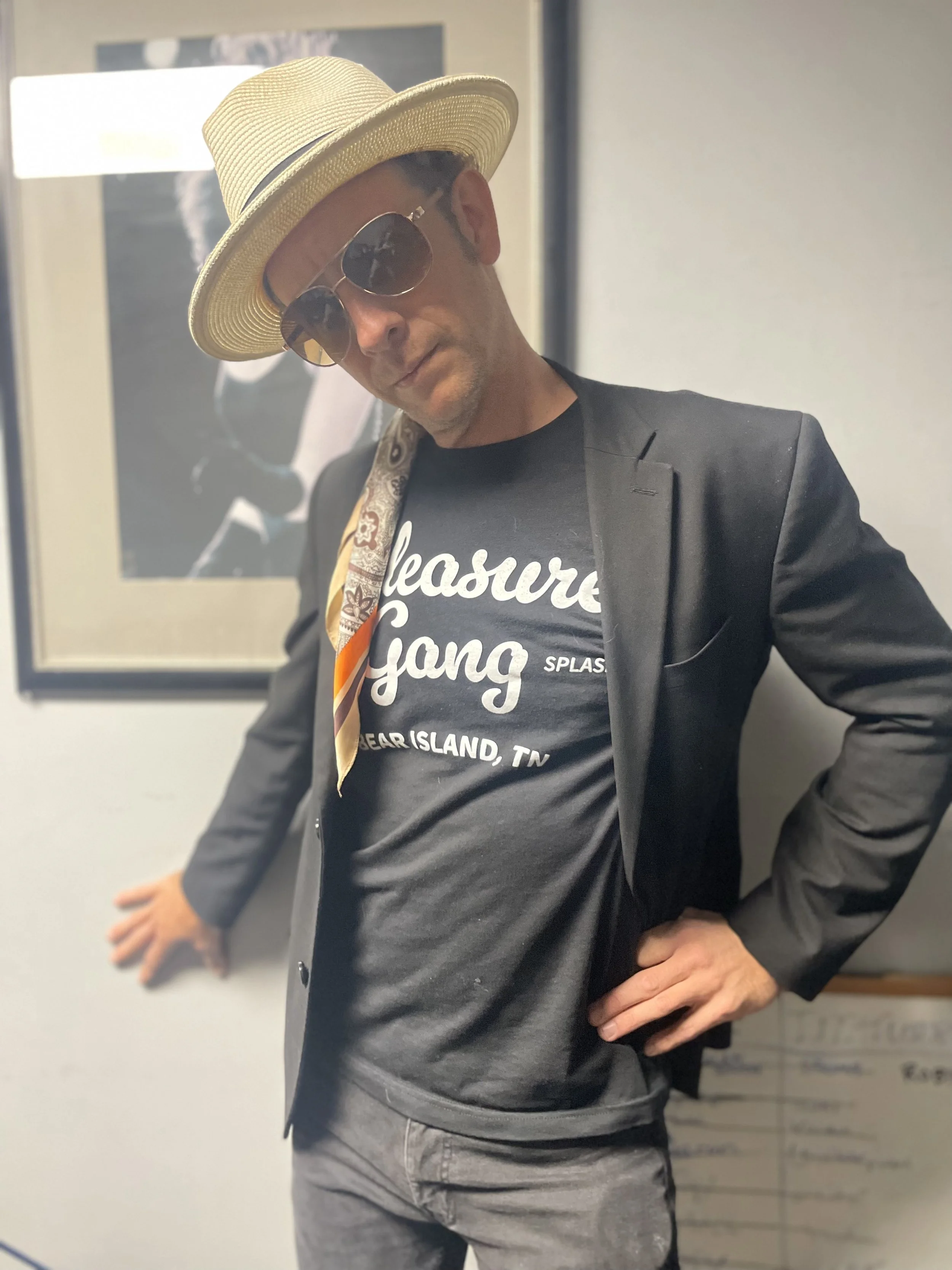 Man wearing a straw hat, sunglasses, a black blazer, a black t-shirt with white text, and a colorful scarf draped over his shoulder, standing indoors with one hand on his hip and the other resting on a door or wall.