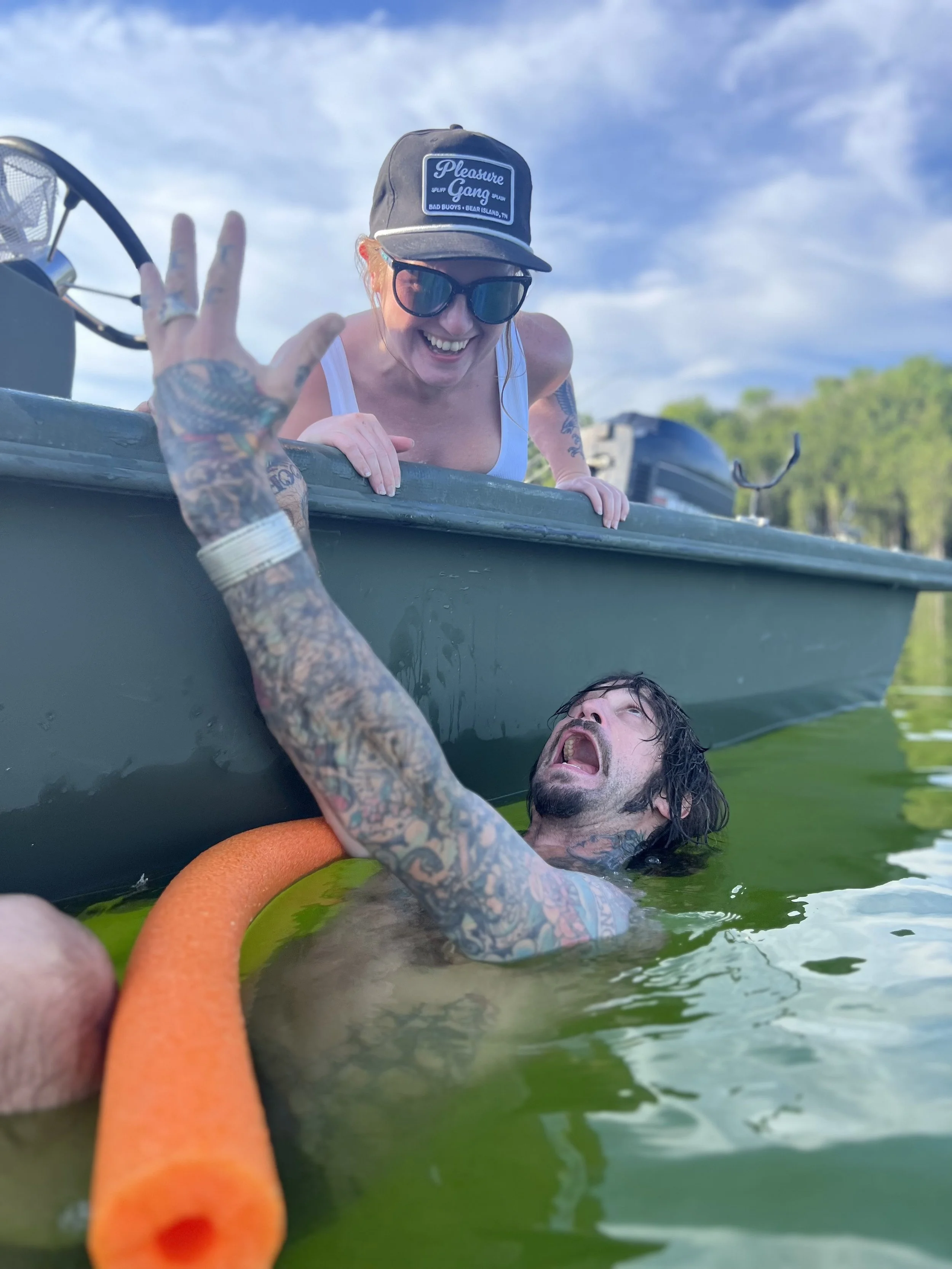 A woman leaning over the side of a boat, smiling and extending her hand to a man in the water, who is holding an orange foam pool noodle near his chest and appearing distressed.
