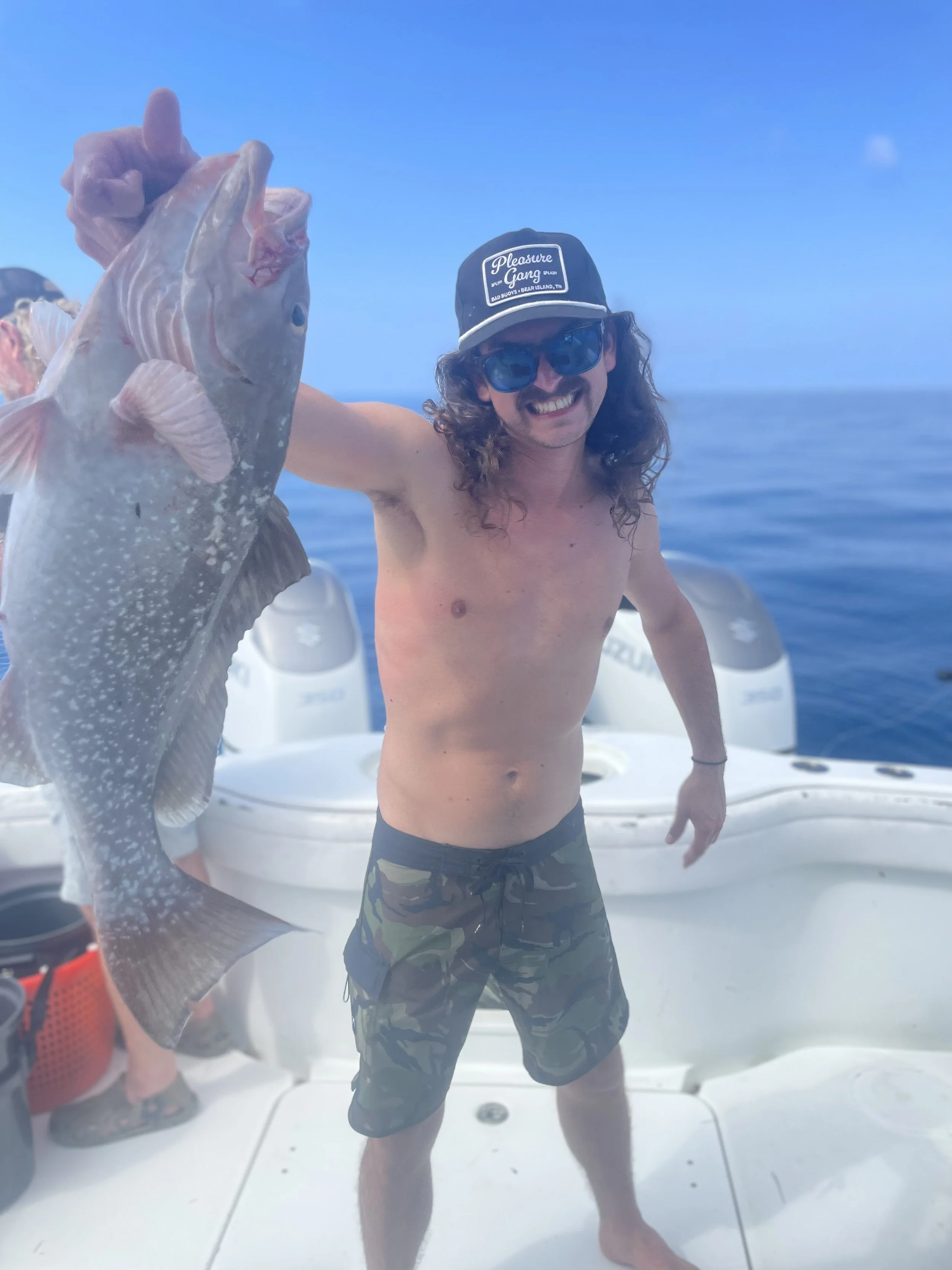 A shirtless man with long curly hair, sunglasses, and a cap holding a large fish on a boat in the ocean.