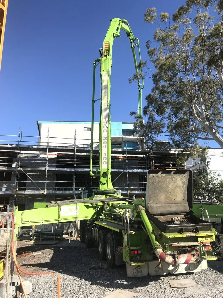 A green concrete pump truck is operating at a construction site with a multi-story building under construction and scaffolding, and trees nearby.