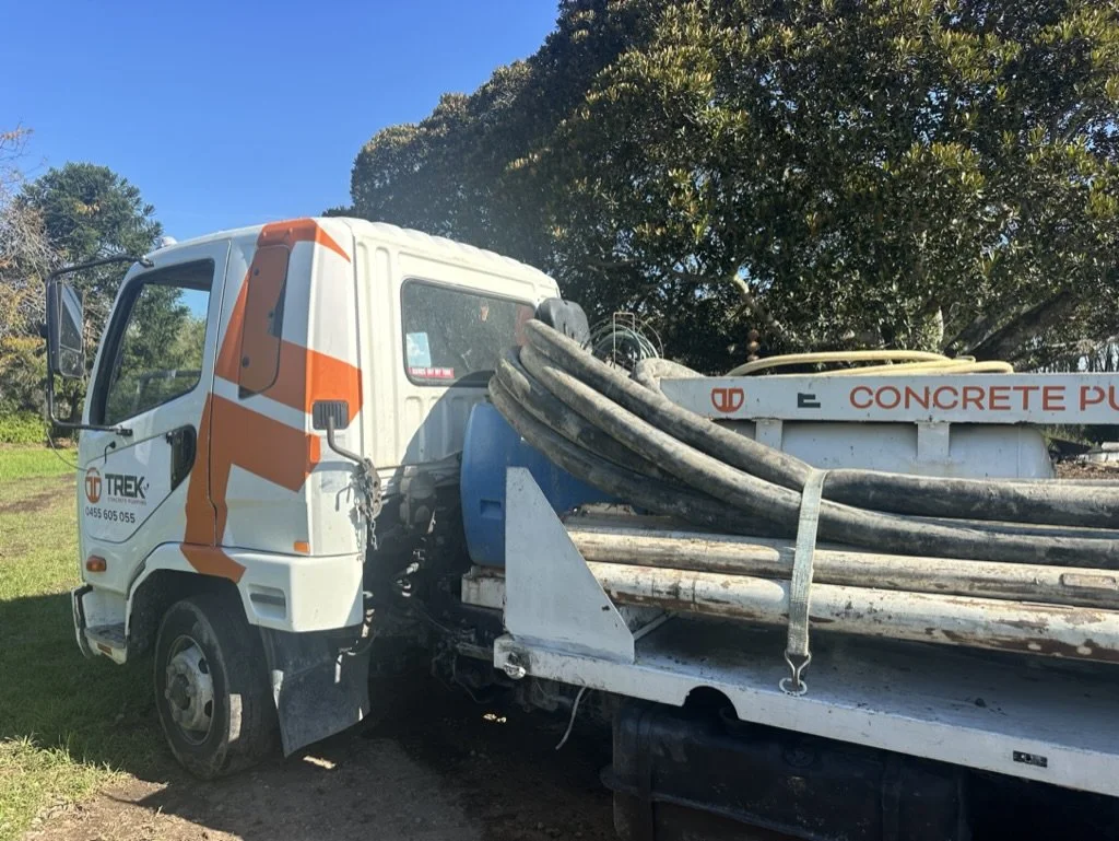 A concrete pump truck parked outdoors on grass with hoses coiled on the side, blue sky, and trees in the background.