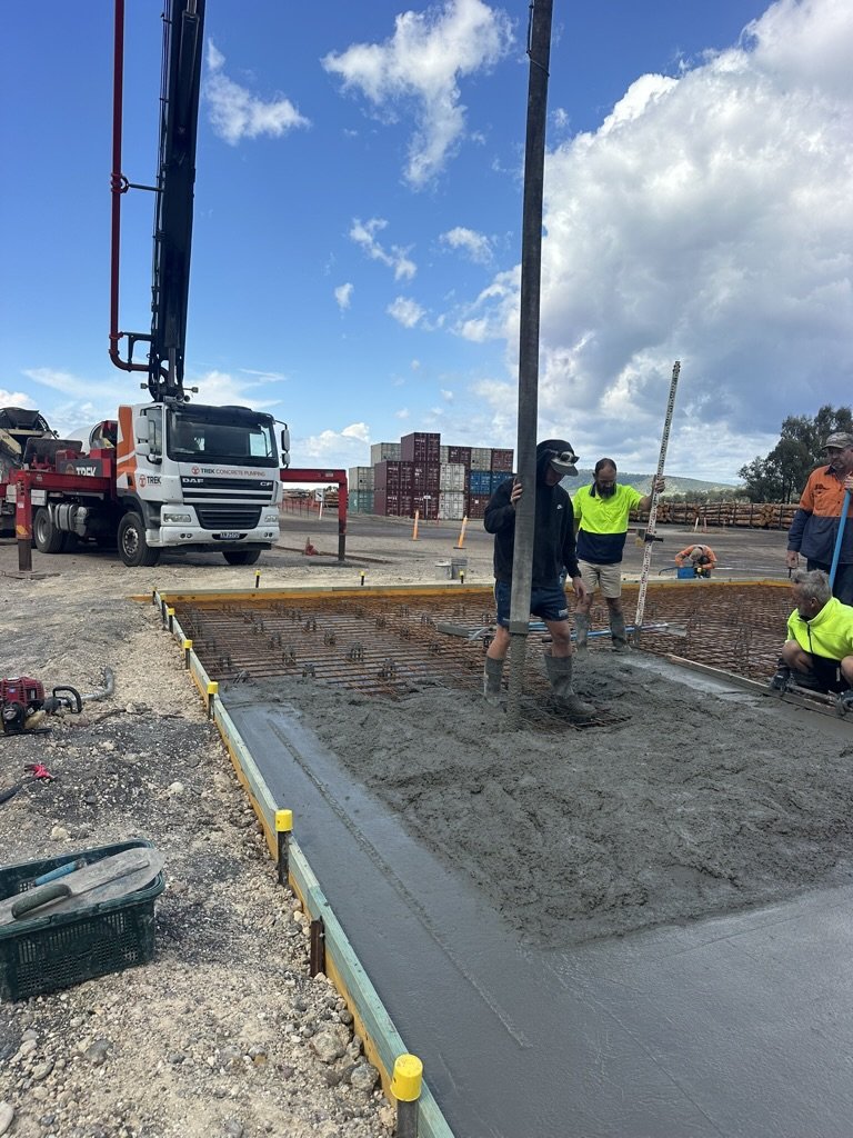Construction workers are pouring and leveling concrete for a building foundation on a construction site, with a crane and shipping containers in the background.