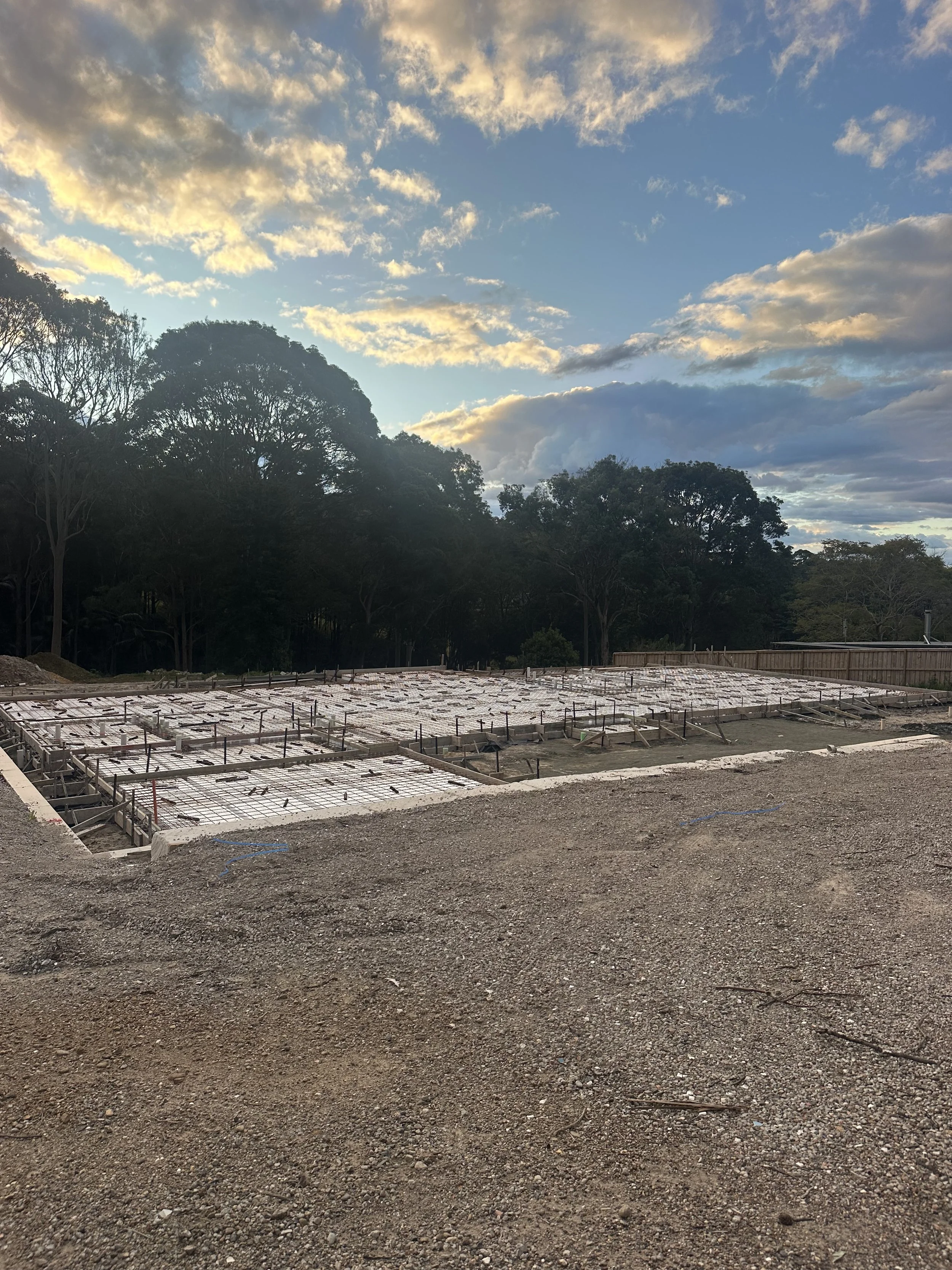 Construction site with concrete forms and rebar, surrounded by dirt and trees under a cloudy sky.