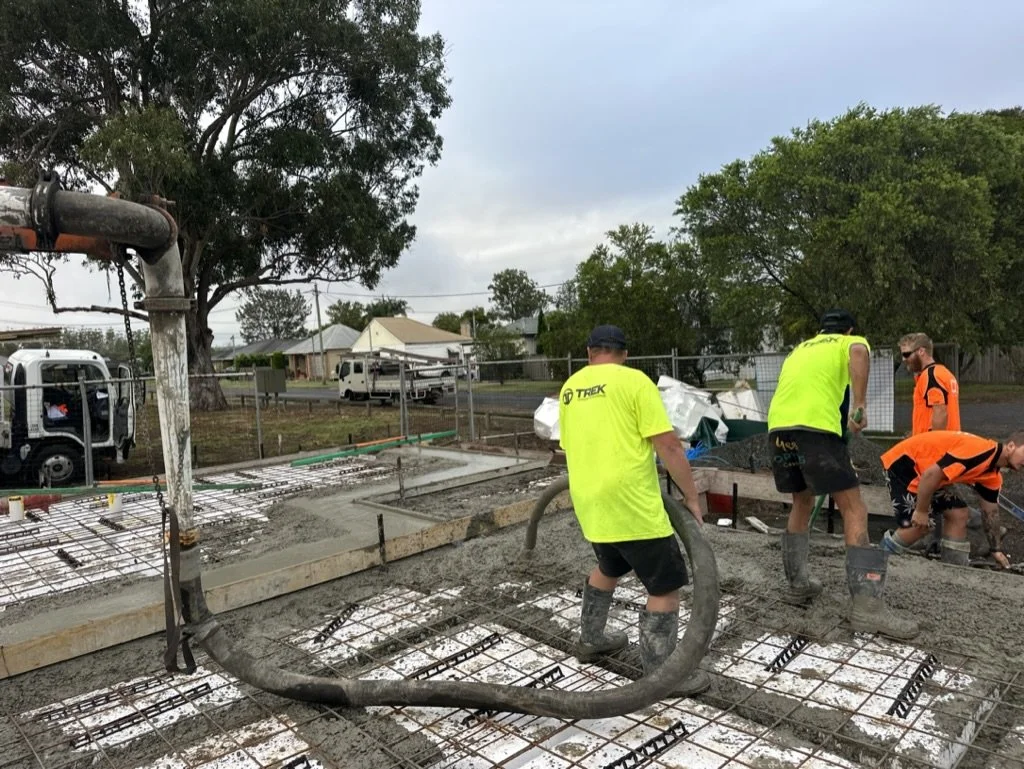 Construction workers pouring concrete on a building foundation with rebar reinforcement, on an outdoor site with trees and vehicles in the background.