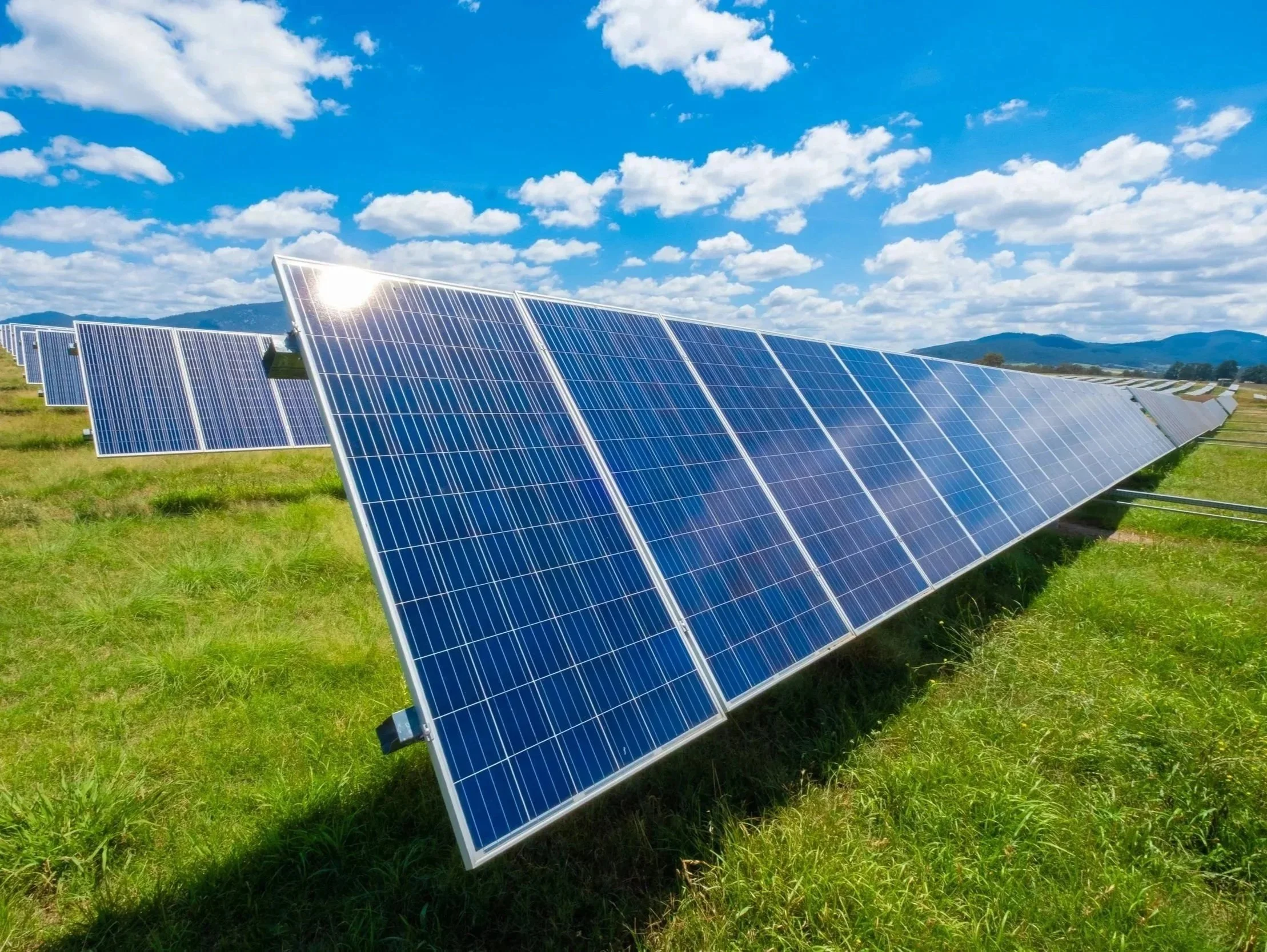 A solar farm with blue solar panels set in a green field under a partly cloudy sky with mountains in the background.
