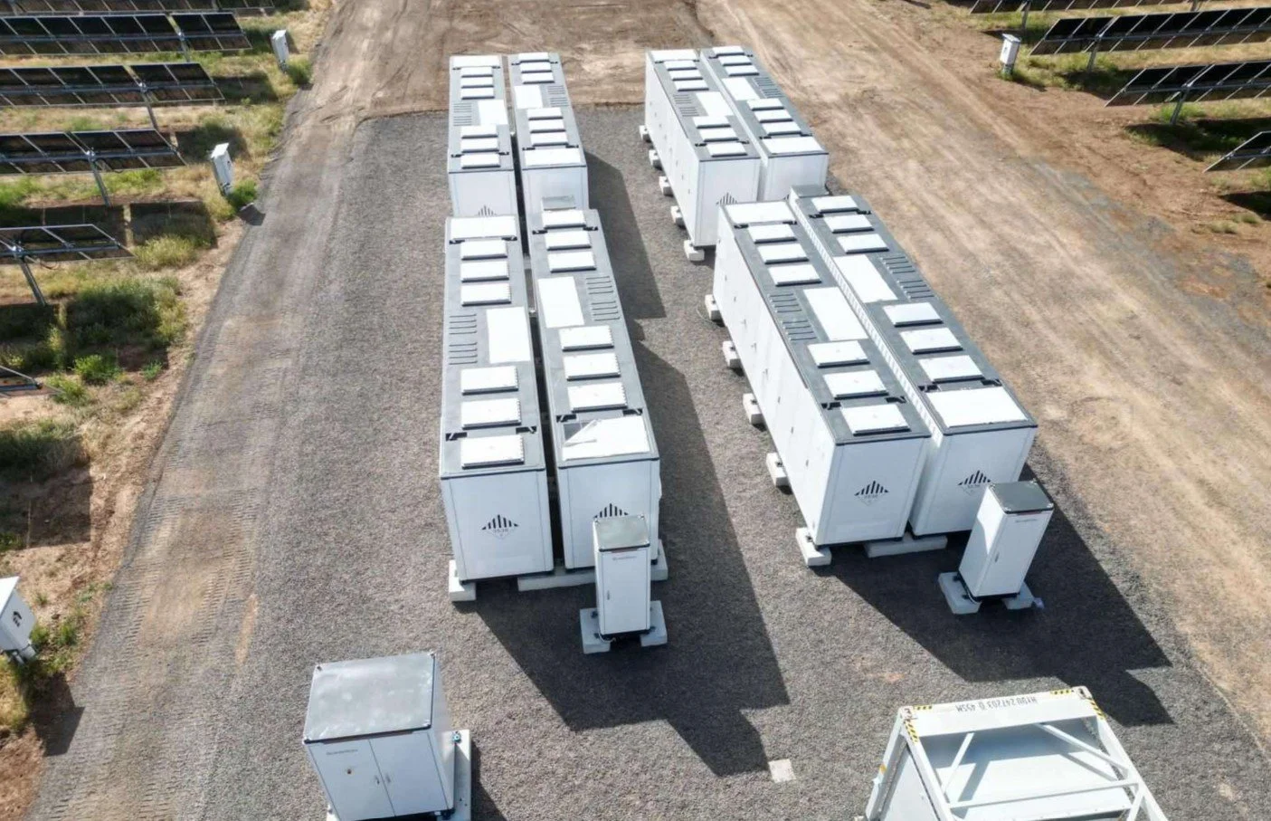 The Hay Solar Farm IFM Battery Energy Storage System - showing array of large white electrical transformers and equipment on a gravel surface in an outdoor facility, with solar panels visible in the background.
