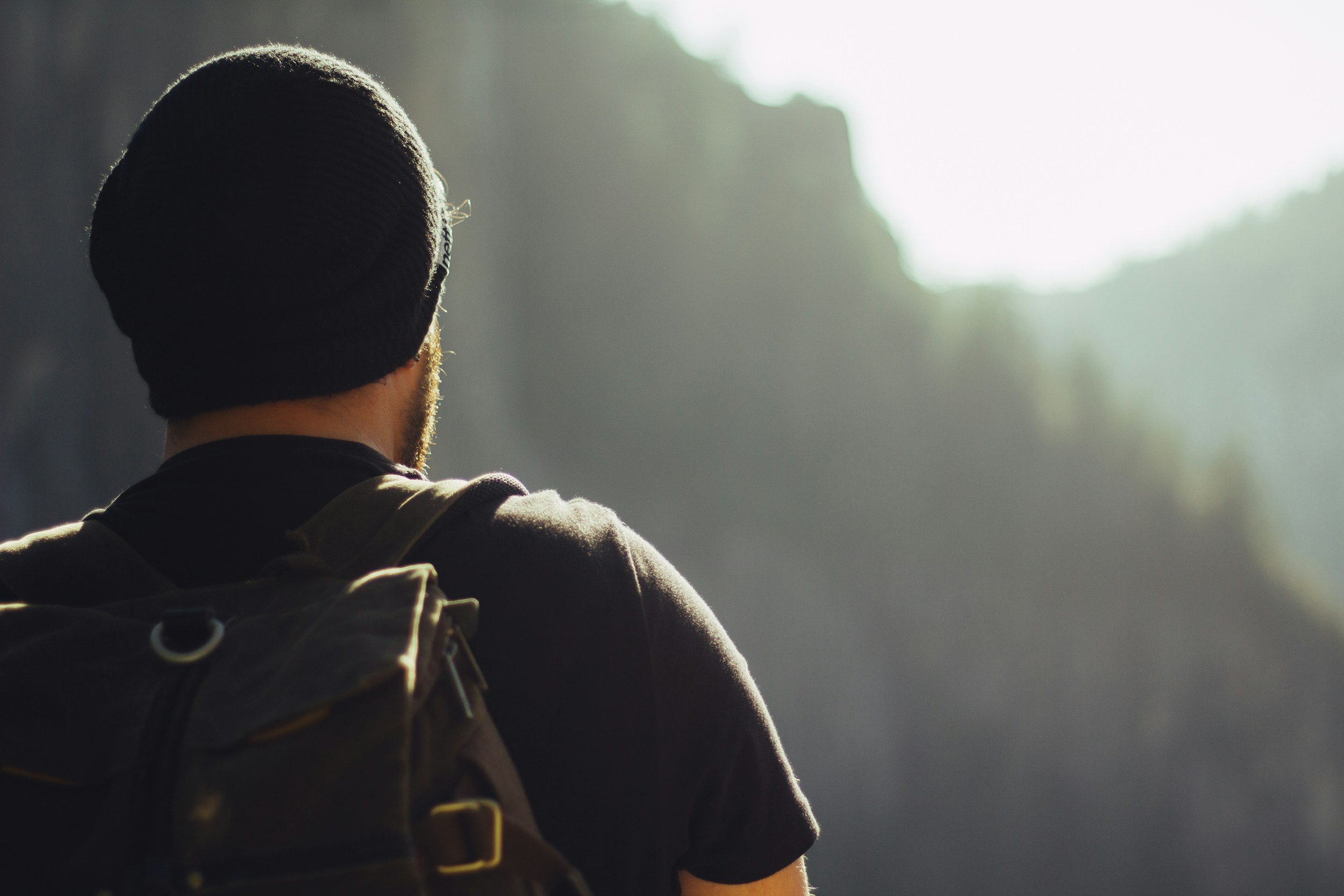 A person wearing a black beanie and glasses, carrying a backpack, looking out towards a bright, foggy landscape with a mountainous area in the distance.