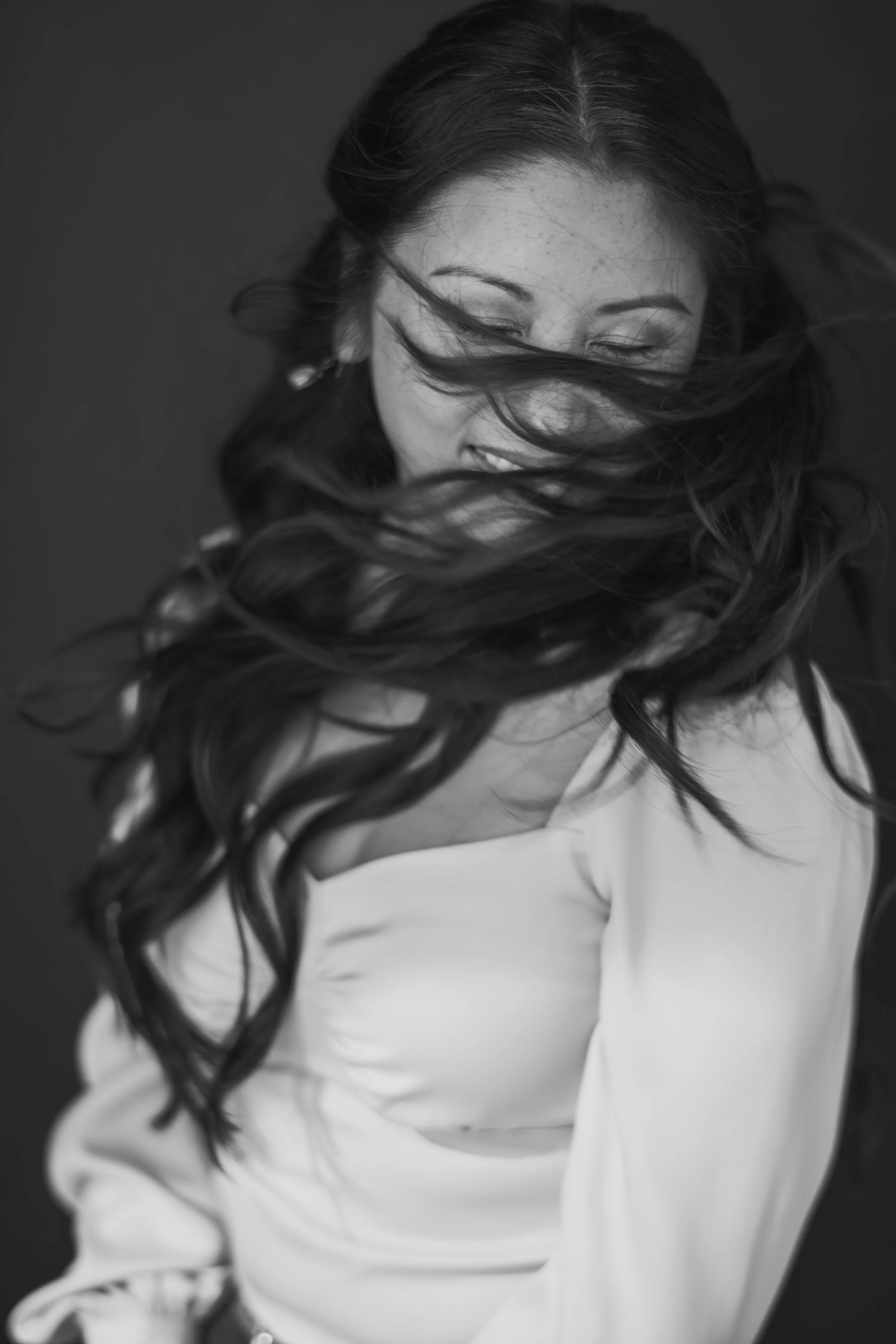 A black-and-white portrait of a woman with long, curly hair, smiling with her eyes closed, wearing a light-colored top.