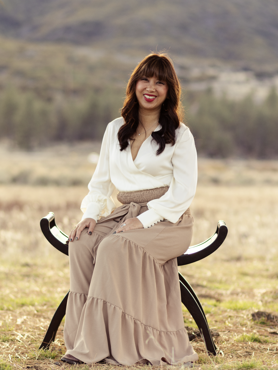 Sonya Lee, executive mentor and author of Channels of Light. A woman sitting outdoors on a bench in a natural setting with mountains and trees in the background. She is smiling and wearing a white blouse and a long beige skirt.