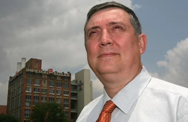 Sonya Lee, executive mentor. John Sheptor, CEO. man with short dark hair and a slight smile, wearing a white shirt and an orange tie, stands outdoors with a cloudy sky in the background. Sign that reads 'IMPERIAL SUGAR' is visible.