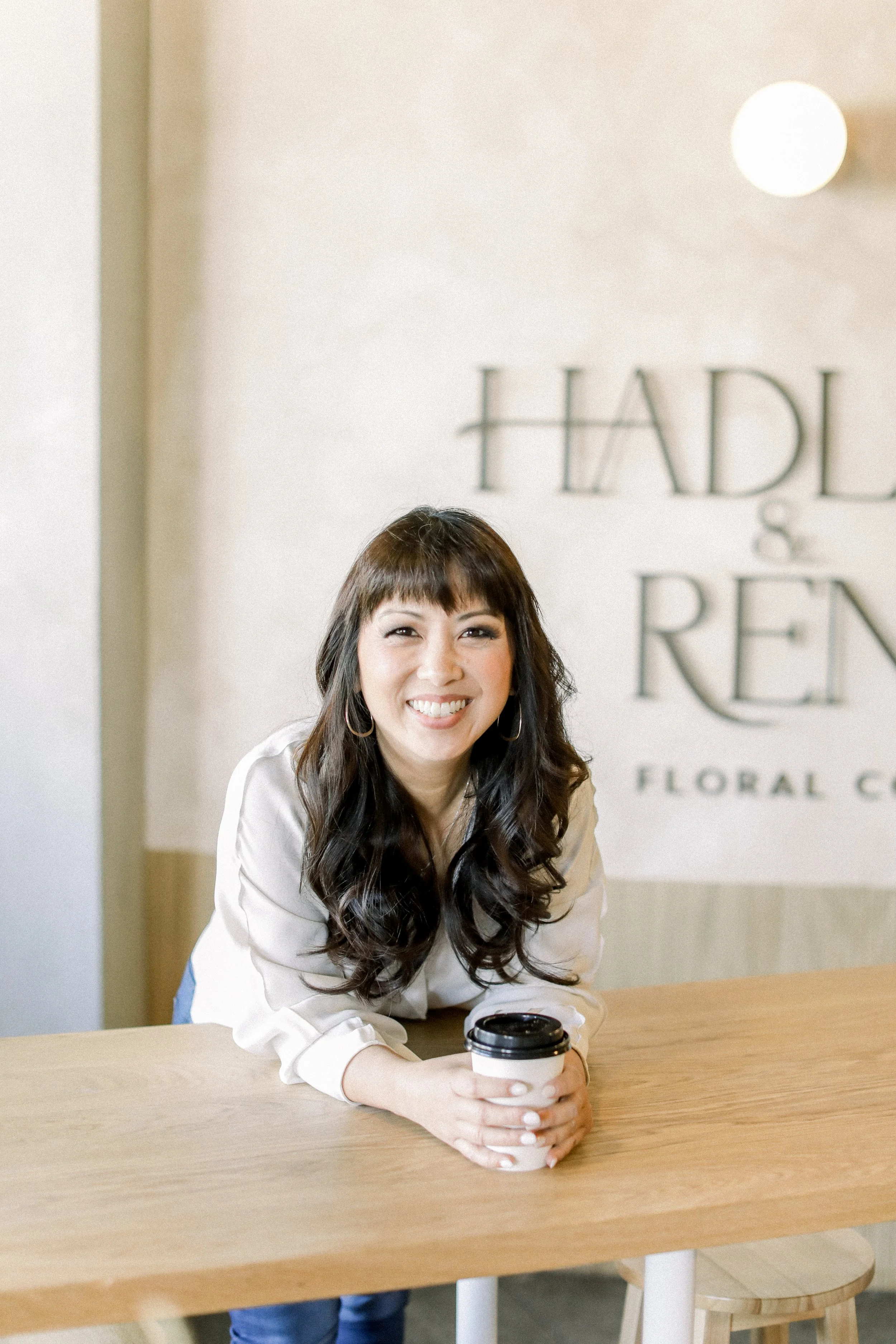 Smiling woman sitting at a wooden table, holding a coffee cup, in a bright modern space with a sign that reads "HADLEY & REEN" in the background.