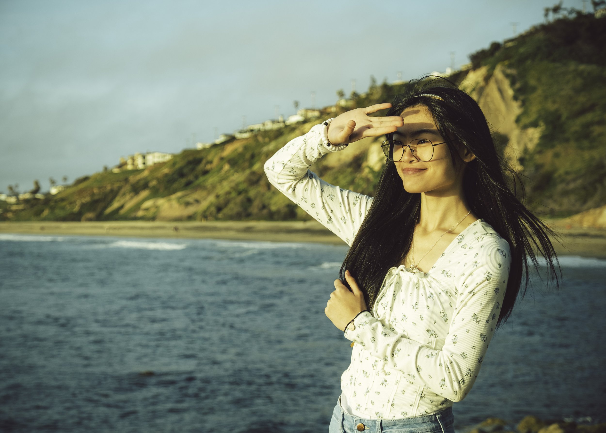 Young woman with long black hair and glasses standing on a beach, shielding her eyes from the sun with her hand, with cliffs and houses in the background.