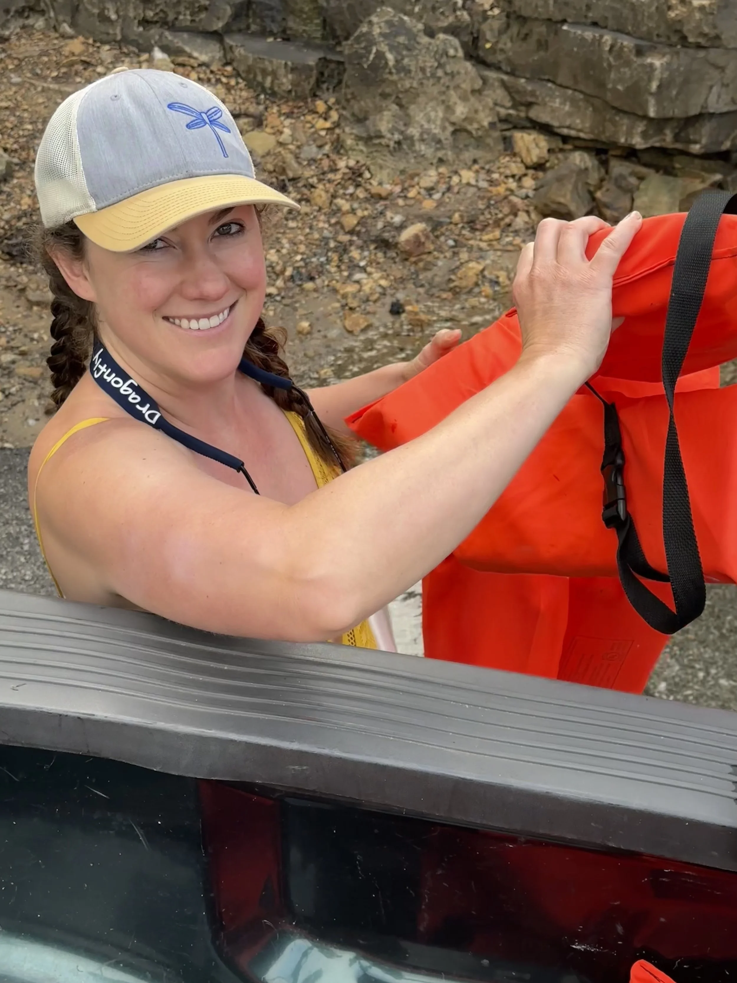 Woman preparing for a kayak adventure at SUP Outfitters in Eureka Springs, holding an orange dry bag near rocky shoreline.