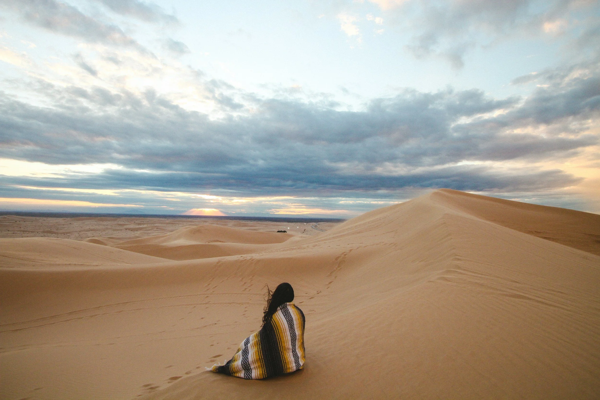 Person sitting on sand dunes in a desert, wrapped in a striped blanket, with clouds and a setting sun in the sky.