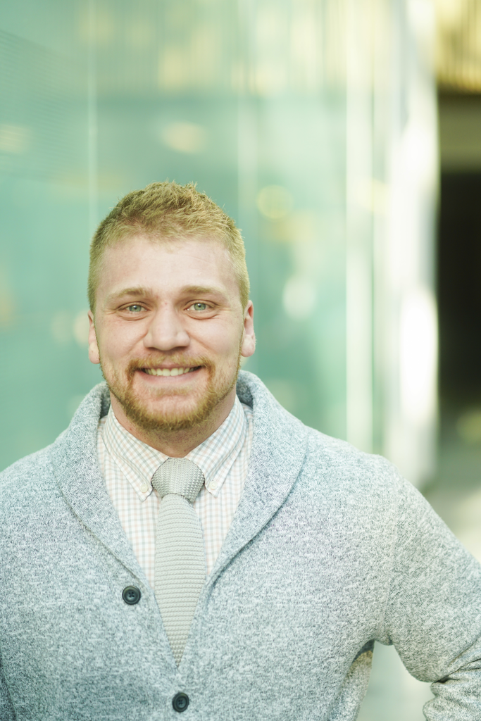 A young man with light hair, beard, and blue eyes smiling at the camera, wearing a light-colored plaid shirt, a light gray tie, and a gray knitted cardigan, standing in a modern building with glass walls and a blurred background.
