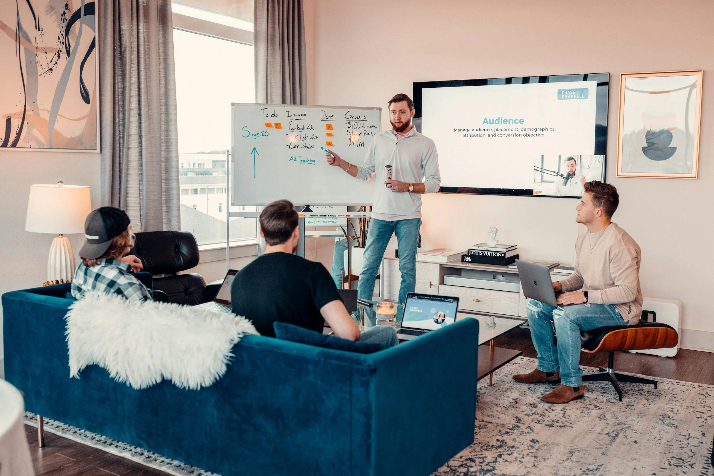 A man giving a presentation in a modern office, with four young adults seated on a blue sofa and chairs, some with laptops, watching him. The whiteboard behind him has notes and goals, and a large screen displays a slide titled 'Audience.'