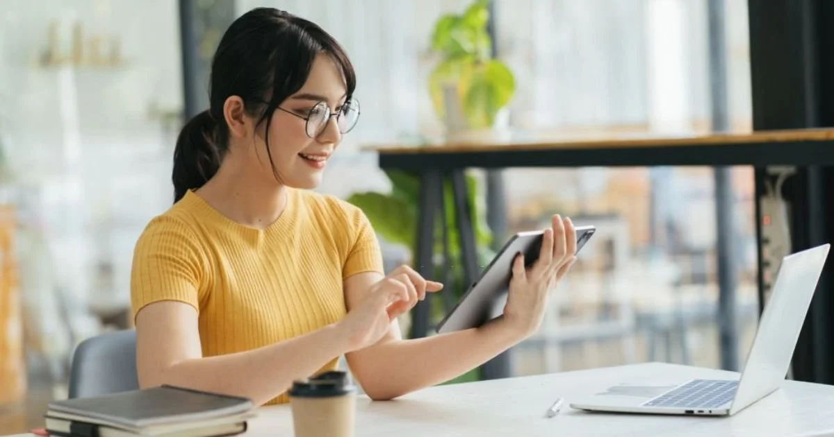 Woman with glasses and a yellow shirt using a tablet at a workspace with a laptop, notebooks, and a coffee cup.