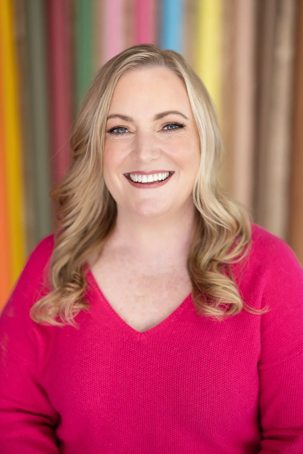 A smiling woman with long wavy blonde hair wearing a pink top, background of vertically striped cloths in various bright colors.