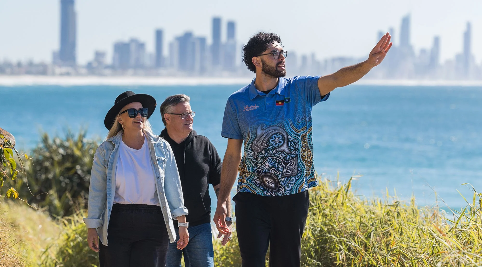 A tour guide leading a group of three people along a coastal area with skyscrapers in the background, water, and greenery.