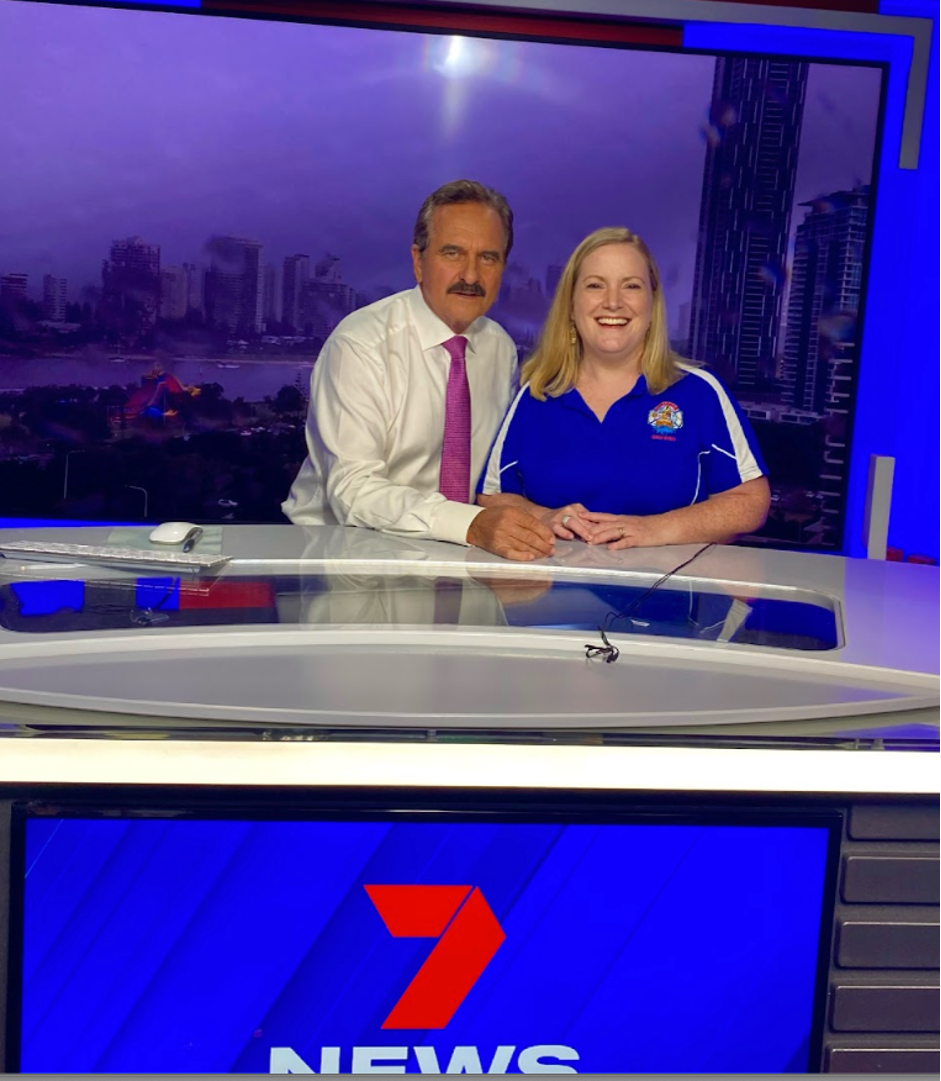 Two news anchors, a man in a white shirt and pink tie, and a woman in a blue shirt with a news logo, sitting in a news studio with a cityscape background and a news desk with a 7 NEWS logo.