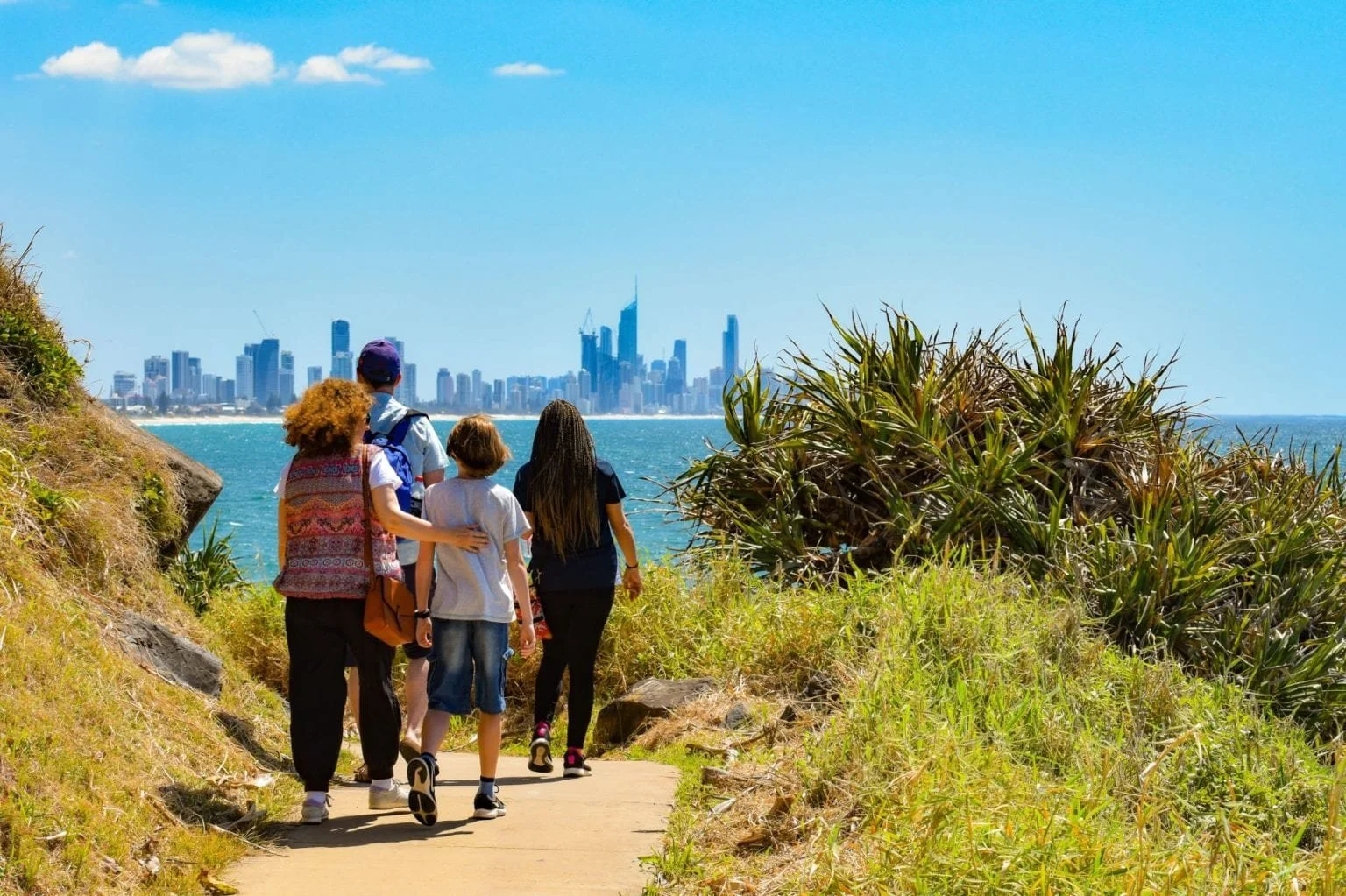A group of five people walking on a trail along a coastal area with a city skyline in the background, including tall skyscrapers, on a sunny day with a blue sky and some clouds.