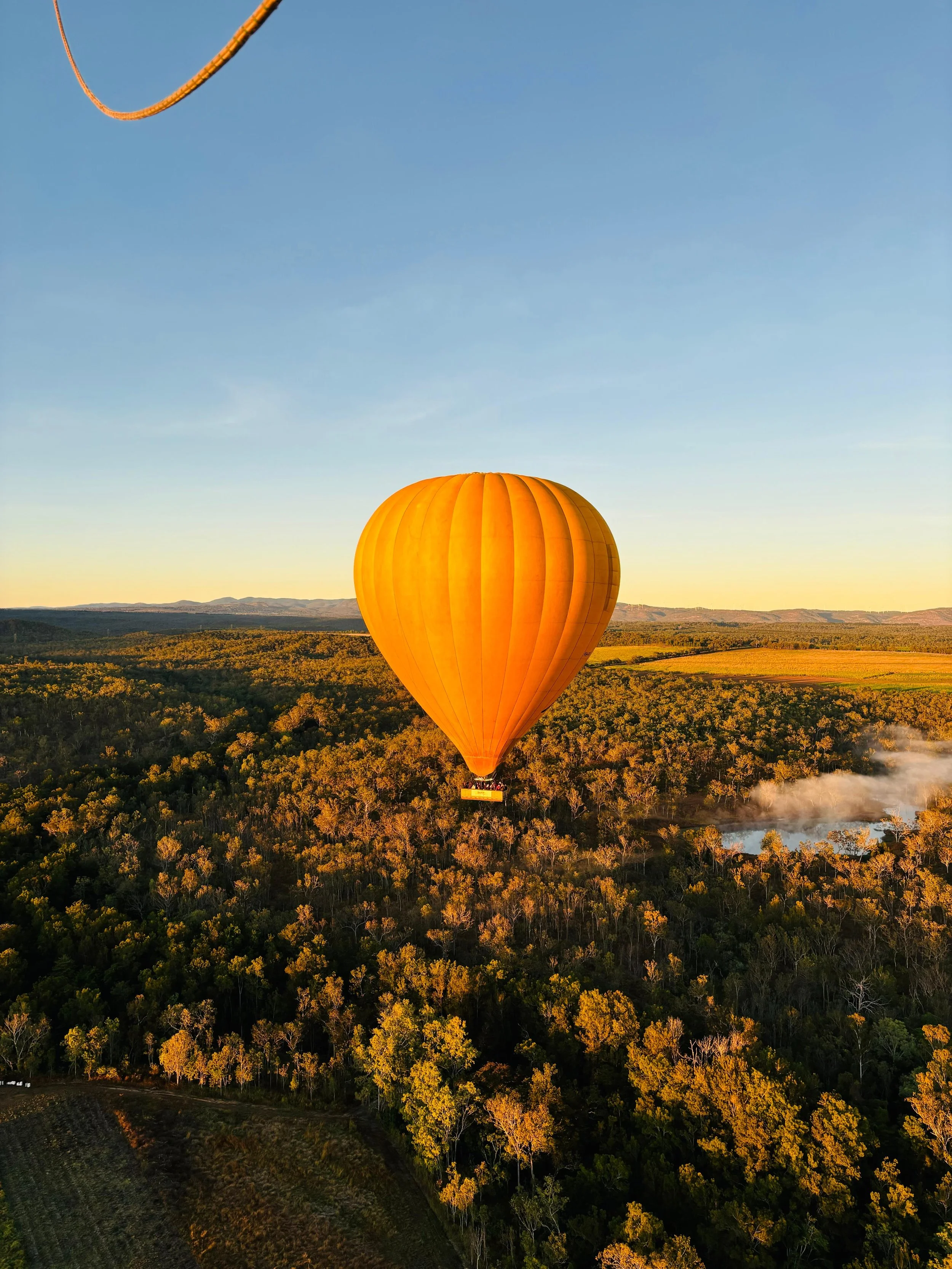 A large orange hot air balloon floating above a forested landscape during sunset with the sky clear and a river in the distance.