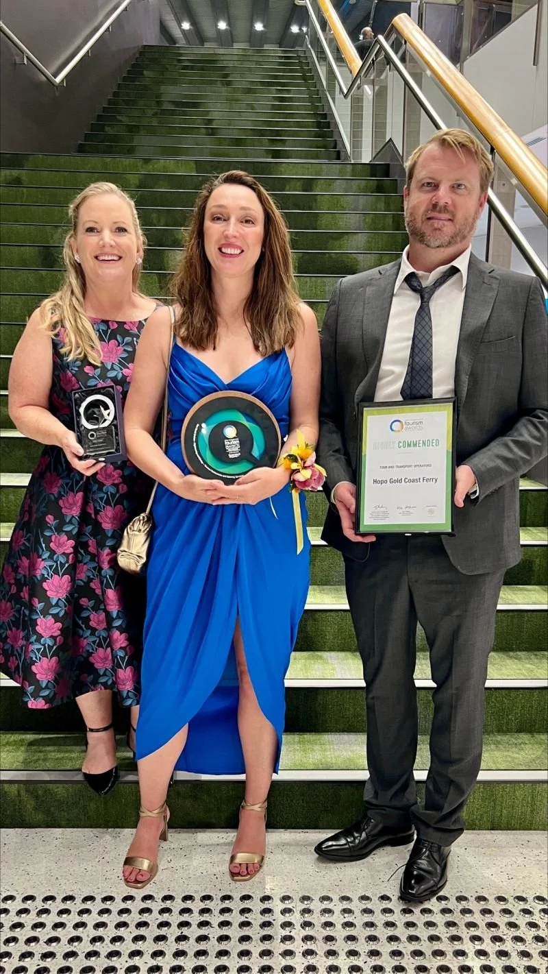 Three people standing on a staircase indoors, holding awards and a framed certificate. The woman in the middle is wearing a blue dress, the woman on the left is wearing a floral dress, and the man on the right is in a gray suit with a white shirt and tie.