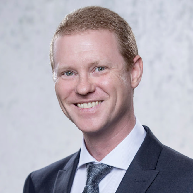 A smiling man with light skin, short red hair, dressed in a dark suit with a white shirt and gray tie, appears to be a professional portrait against a plain light background.