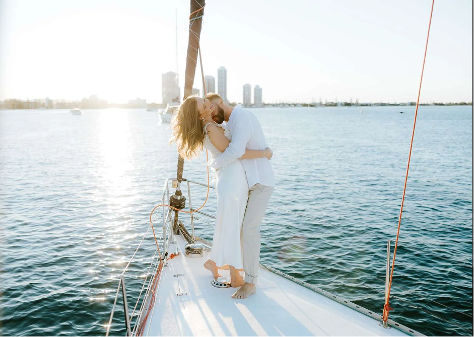A couple hugging on a sailboat at sunset on the water with city skyline in the background.