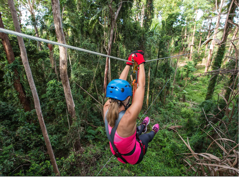 Woman ziplining through a dense green forest wearing a blue helmet, pink top, and safety harness.