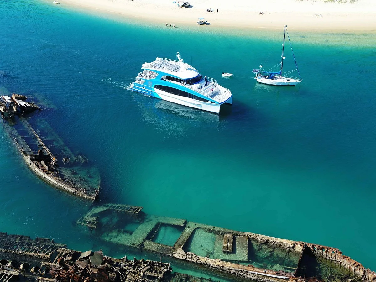 A large white and blue yacht sailing near a partially submerged, rusted sunken shipwreck in turquoise waters near a sandy beach with small boats.