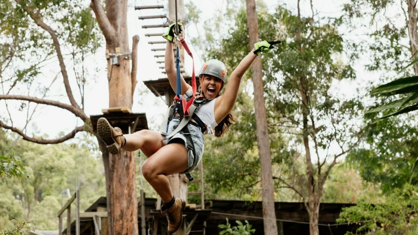 Woman in helmet and harness swinging on zip line through a forested area with trees and a treehouse in the background.