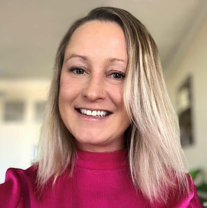 A woman with blonde hair, blue eyes, and a nose piercing smiling, wearing a pink top, indoors.
