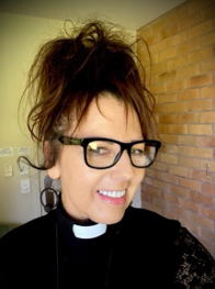 A woman dressed as a priest with black glasses, earrings, and a black clerical shirt with a white collar stands outdoors in front of greenery and a white lattice fence, smiling slightly and making a grimace.
