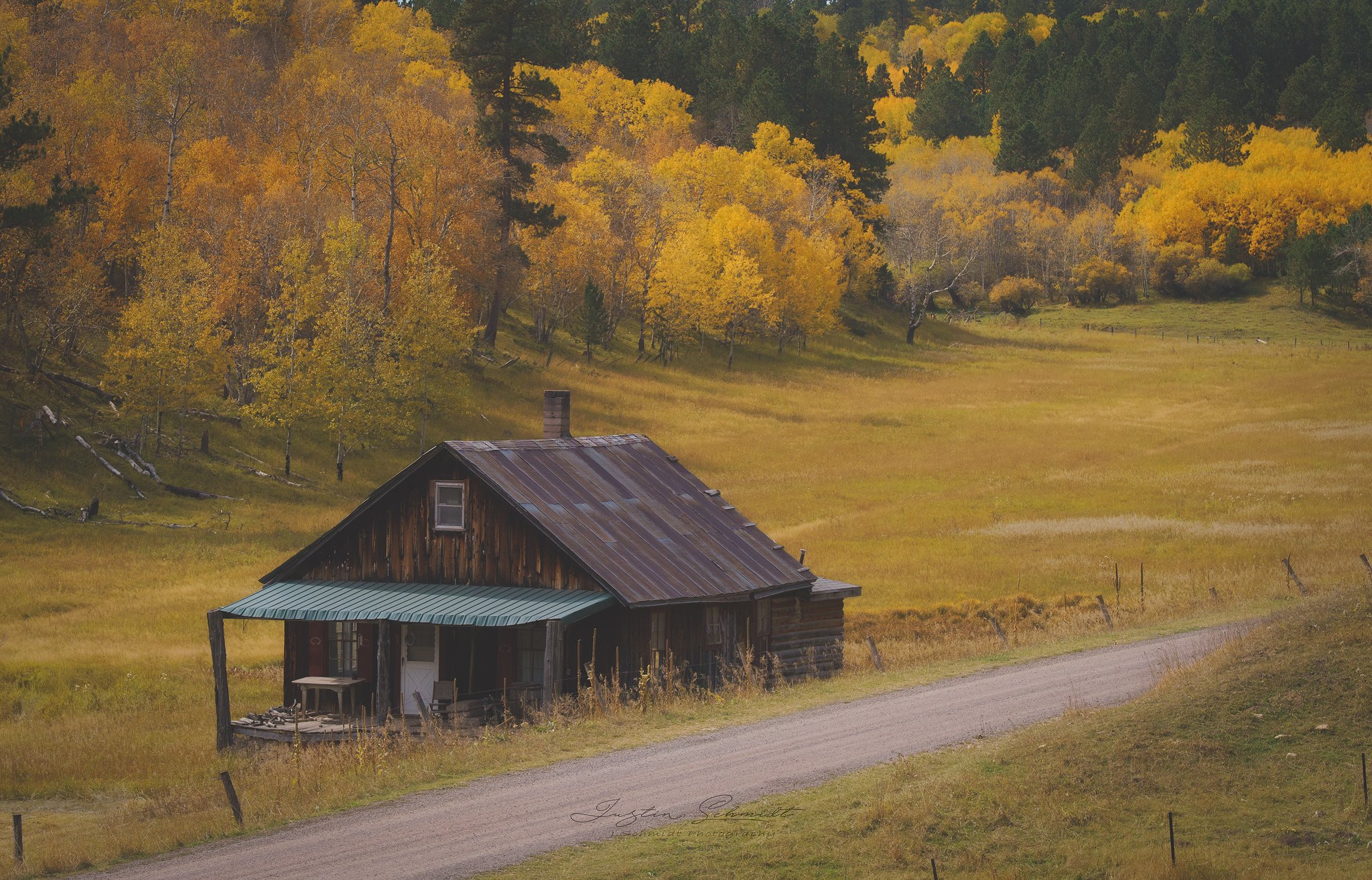An old rustic wooden house with a metal roof, sitting on a grassy field next to a dirt road, with a backdrop of yellow and green trees in a hilly landscape during autumn.