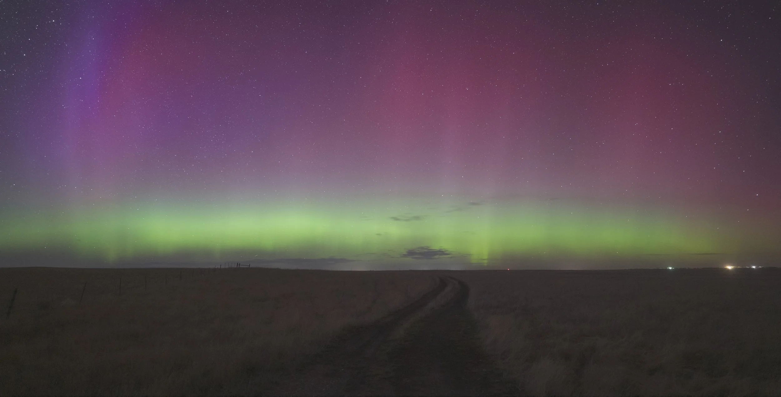 Night sky with colorful northern lights, stars, and a dark landscape with a dirt road and a few small lights on the horizon.