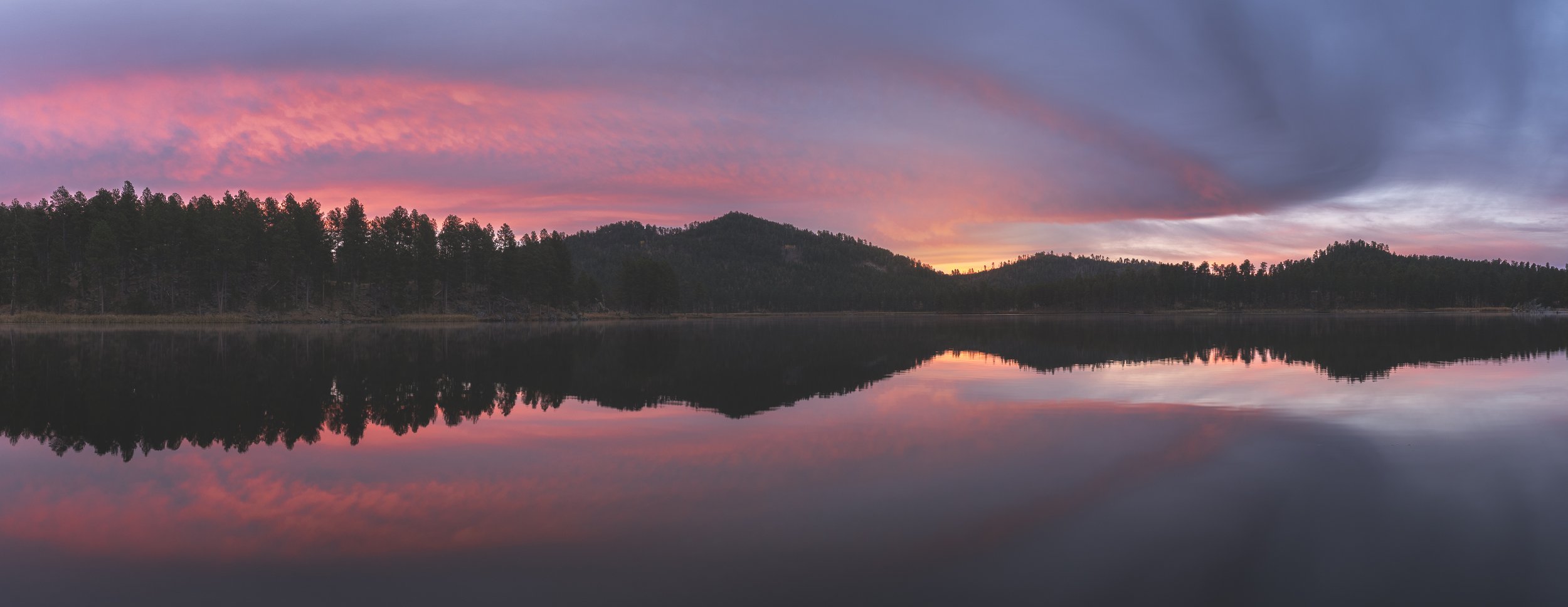 A serene lake reflecting a colorful sunset sky with pink and purple clouds, surrounded by tree-covered hills.