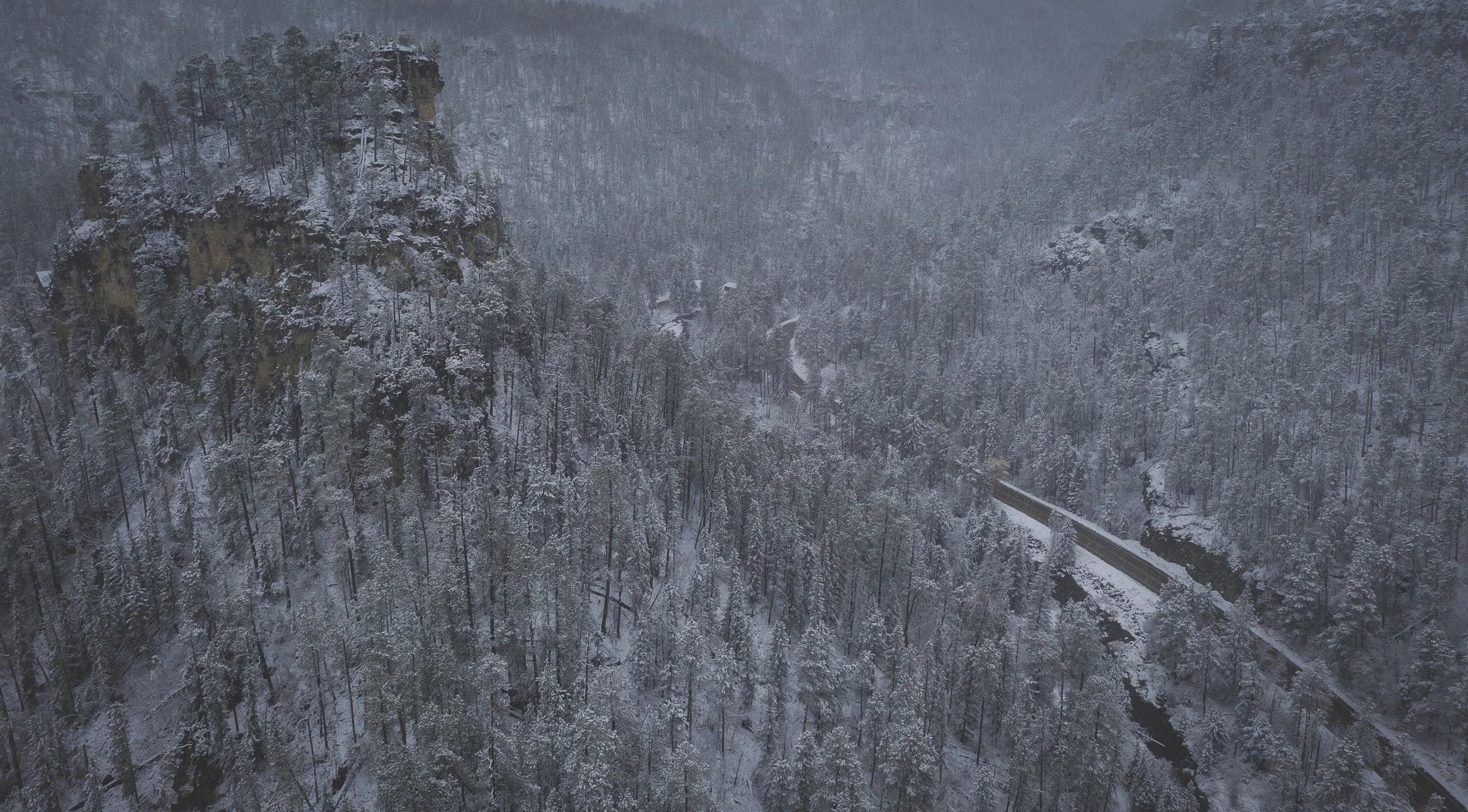 An aerial view of a snow-covered mountain forest with a winding road and a rocky cliff on the left side.